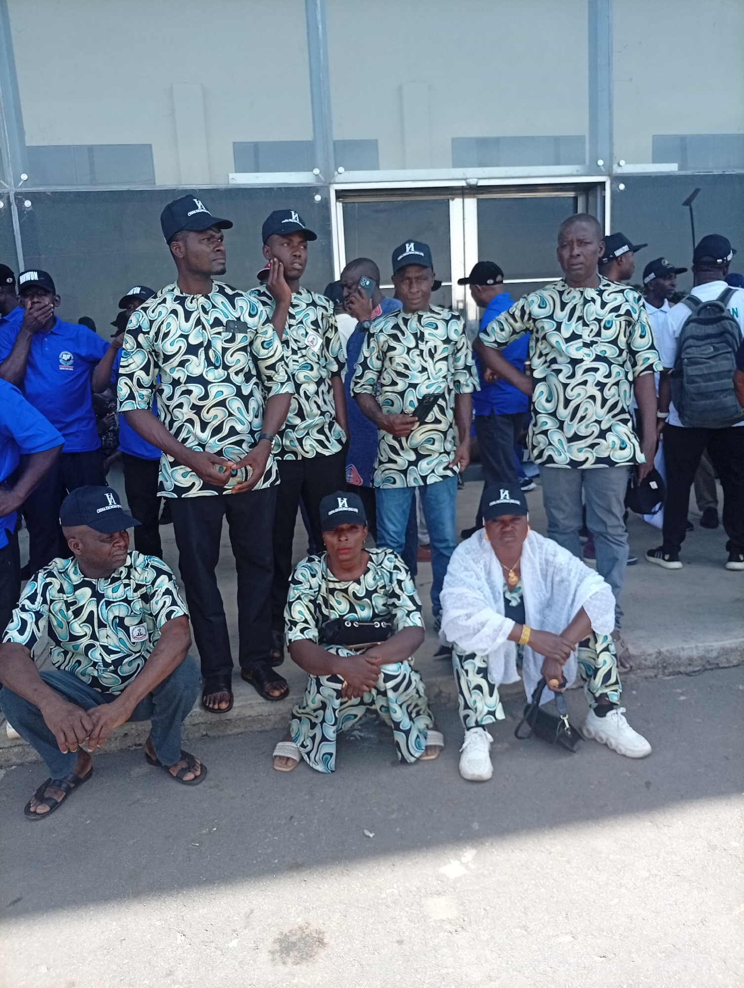 In Unguwar Sarki, Nigeria, a group of nine people, most of whom are wearing identical patterned shirts and black baseball caps, are situated outdoors in front of a modern building with glass paneling. Five individuals are standing, while four are squatting or sitting on a low ledge. The clothing suggests a uniform or affiliation. Other people are partially visible in the background. The ground appears to be paved, and the lighting suggests it is daytime.
