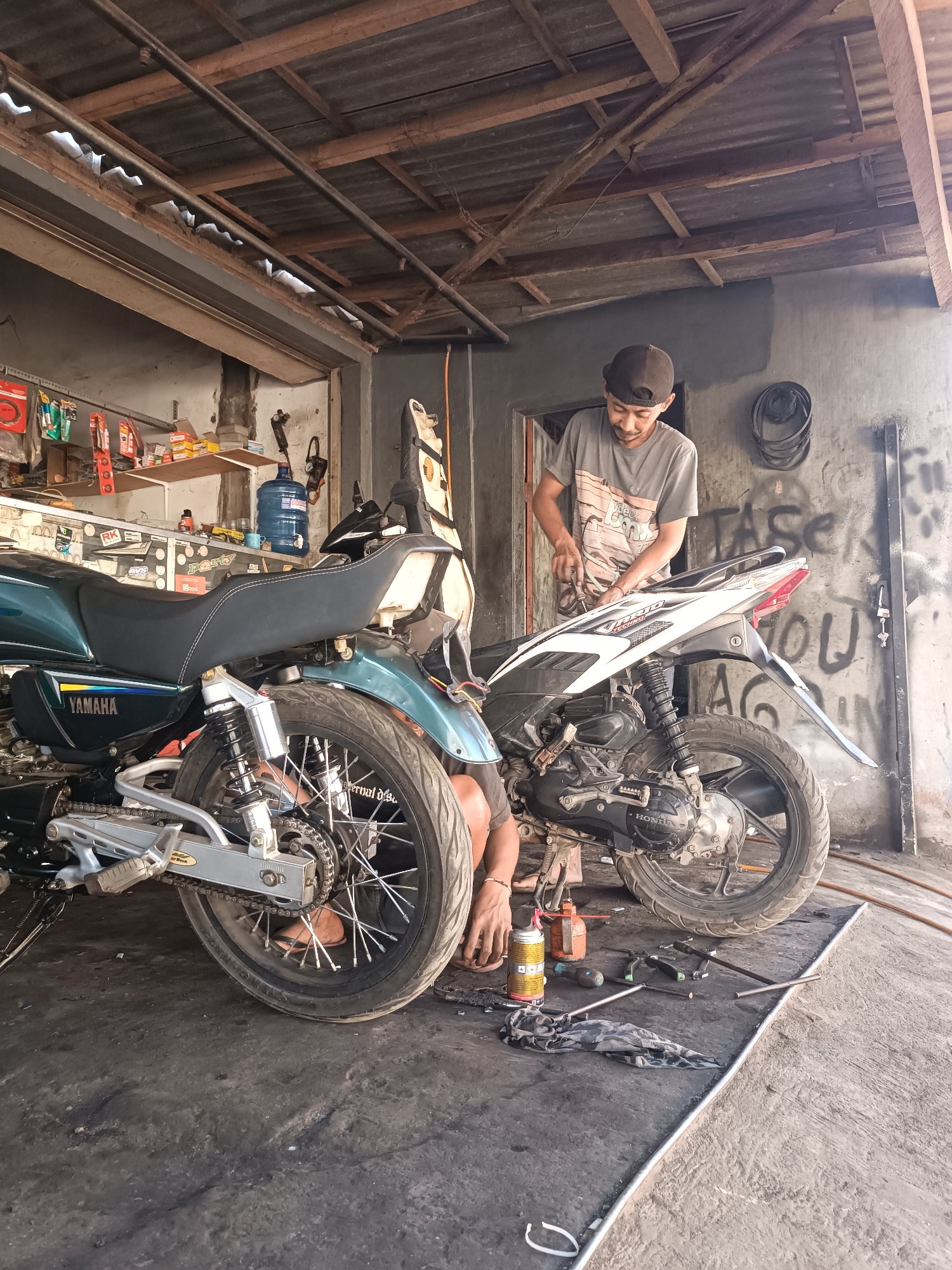 The image depicts a motorcycle repair shop in Brongkal, Indonesia. A man in a grey t-shirt and a baseball cap is working on a white motorcycle. Another person's legs, wearing flip-flops, are visible on the left side of the frame, appearing to be working on a green Yamaha motorcycle. The floor is concrete and stained with oil and dirt. Various tools, including wrenches and a spray can, are scattered around the motorcycles. Shelves with spare parts and supplies are visible in the background. The roof is made of corrugated metal supported by wooden beams. The wall behind the motorcycles is concrete with graffiti.