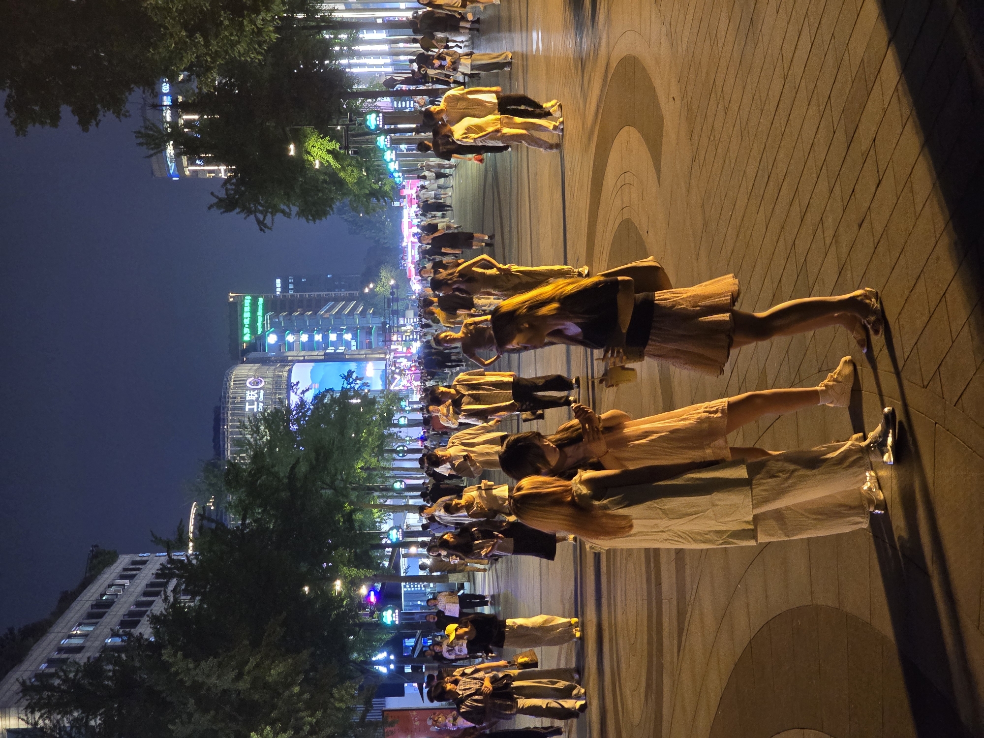 This image depicts a wide, illuminated pedestrian street at night within Xihu District, China. Numerous individuals are present, primarily appearing to be young adults and adults.
In the immediate foreground, three women are standing and interacting. The woman on the left has long light-colored hair and wears a loose-fitting, light-colored long-sleeved top over wide-leg light-colored pants, paired with reflective silver-colored shoes. She is oriented slightly to the right of the frame. The central woman has long dark hair and wears a light peach- or cream-colored short dress with light-colored sneakers; she is holding and looking at a smartphone. The woman on the right has long dark hair, wears a dark short-sleeved top, a patterned light-colored skirt, and open-toed shoes, and is holding a yellow cup while looking at the central woman's smartphone.
Further down the street, a crowd of people is walking, standing, and conversing. Their attire is casual, consisting of various shirts, pants, and dresses. One individual in the mid-left wears a yellow baseball cap. The street pavement features a pattern of dark and light-colored tiles arranged in linear and curved designs.
The street is flanked by multiple-story buildings and mature trees. The trees are illuminated from below, casting a green glow on their foliage. Various streetlights with both white and colored (green, blue) light sources are visible. Buildings along the street feature illuminated windows, large digital display screens showing various graphics or advertisements, and prominent signage. One building, identifiable in the upper left section of the image, displays Chinese characters that translate to "Gōnglián" (工联) alongside the English text "City Center". Artificial light sources create distinct shadows of people and objects on the pavement. The sky above is dark, indicating nighttime conditions.
