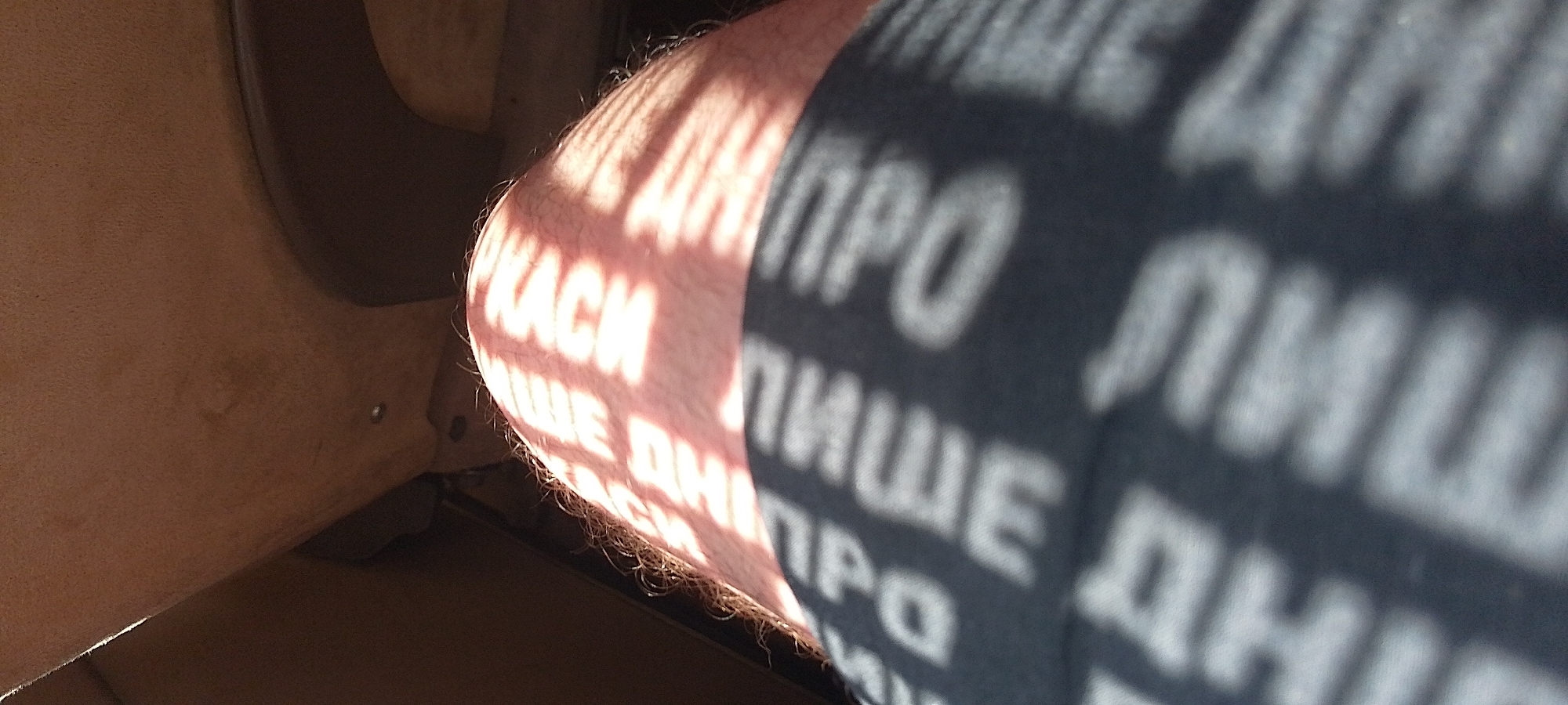A close-up, angled shot shows a person's leg and thigh, covered by a dark fabric band with white text. The text on the fabric band is difficult to read due to blurriness and shadowing, but appears to be repeated words such as "TPQ" and "MAUE" in a stylized font. A pattern of sunlight and shadow falls across the skin of the upper leg, creating horizontal stripes of light. The background is out of focus, appearing to be the interior of a vehicle with brown upholstery. There is no visible indication of the city of Cherkasy, Ukraine, or any specific urban environment. The image focuses solely on the leg and the fabric band.
