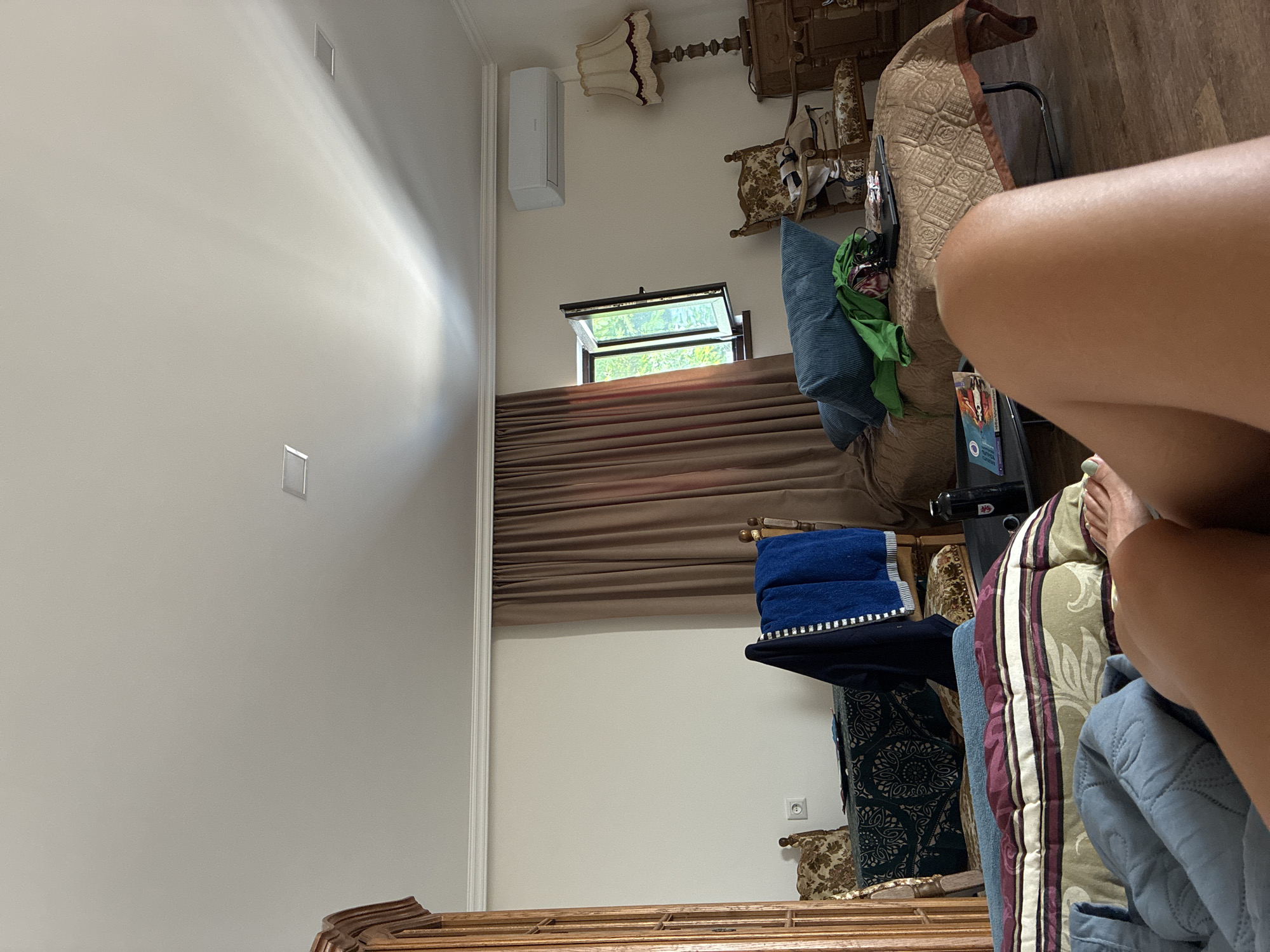 The image depicts an indoor scene, likely a bedroom or living area, in Surami, Georgia. The perspective is from a low angle, looking towards a bed where a person's legs are visible. The room contains a wooden wardrobe on the left, a table with two chairs covered in patterned fabric, and a blue towel draped over one chair. In the background, there is a window with open shutters revealing greenery outside, a brown curtain, and an air conditioning unit mounted on the wall. A lamp with a patterned shade sits on a wooden dresser. There is also a daybed with pillows and a green garment, and a small table with books and a water bottle. The overall impression is of a lived-in space.