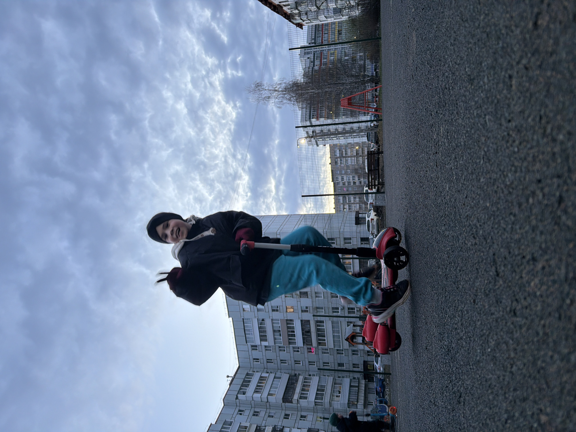 An outdoor shot captures a person, likely a child or young adult, enthusiastically posing on a red scooter on a paved surface. The subject is positioned in the left-center, looking towards the camera with a wide smile and flashing a peace sign with their left hand. They are dressed in a black beanie, a black padded jacket with white lining visible on the hood, and bright turquoise sweatpants, paired with dark sneakers.
The background features a typical urban environment. Behind the person and scooter, a wire mesh fence extends across the mid-ground. Beyond the fence, several multi-story residential buildings, consistent with post-Soviet architecture, dominate the skyline. A bare tree, indicative of autumn or winter, stands behind the fence, along with a red playground structure. In the distance, cars are parked, and a park bench is visible. Another person, partially visible in the bottom right corner, wears a dark jacket and a green beanie.
The sky is heavily overcast with dense, grey clouds, suggesting either a cloudy day or late afternoon. The overall lighting is subdued. The paved ground, likely asphalt, occupies the foreground and right side of the image. The red scooter has "Y*S" visible on its front. The subject's joyful expression and peace sign convey a happy and playful mood. The scene aligns with the context of Chaikovsky, Russia.