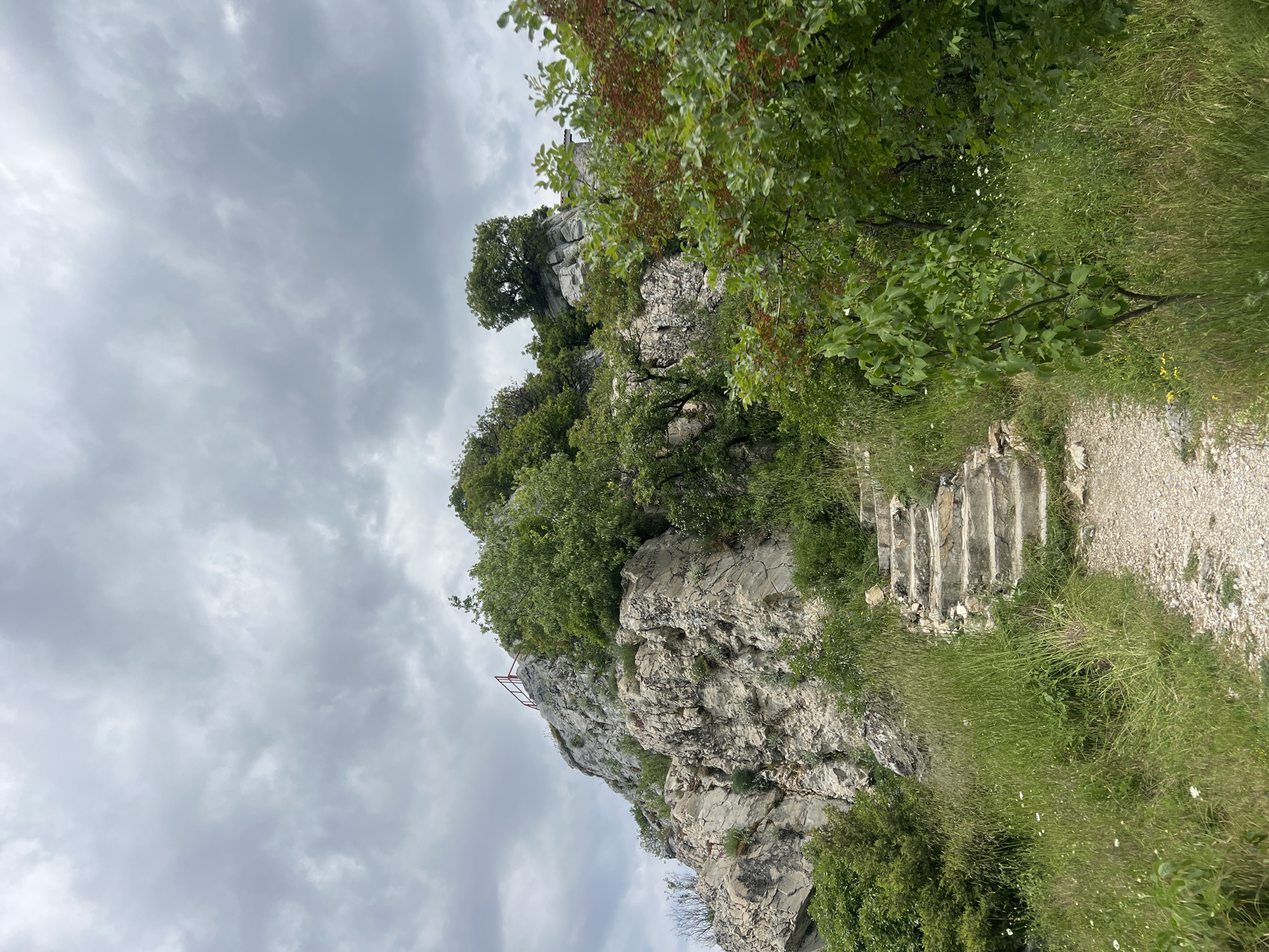 The image shows a rocky outcrop in Asenovgrad, Bulgaria. The outcrop is covered in vegetation, including trees and shrubs. A set of stone steps leads up the side of the outcrop to a gravel path. A red metal railing can be seen on top of the outcrop. The sky is overcast.