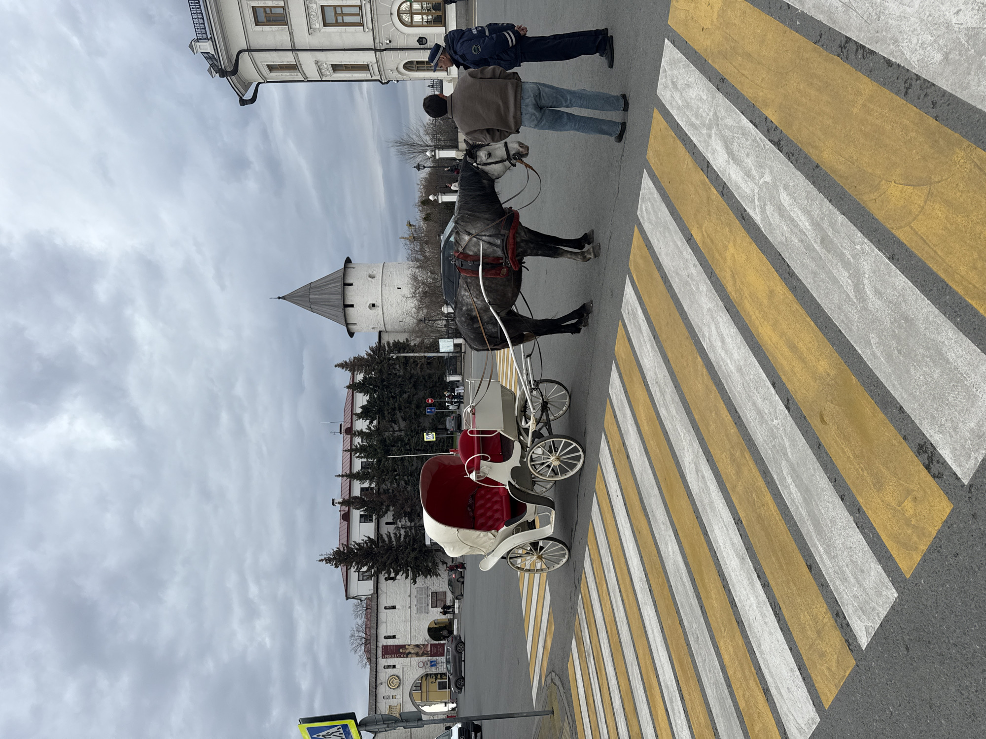 The image shows a horse-drawn carriage in Kazan, Russia. The carriage is white with red seats. The horse is gray. Two men stand near the horse; one is wearing a brown jacket and jeans, while the other is wearing a dark blue uniform and hat. They are positioned on a crosswalk with yellow and white stripes. In the background, there is a building with a tower. The sky is overcast.