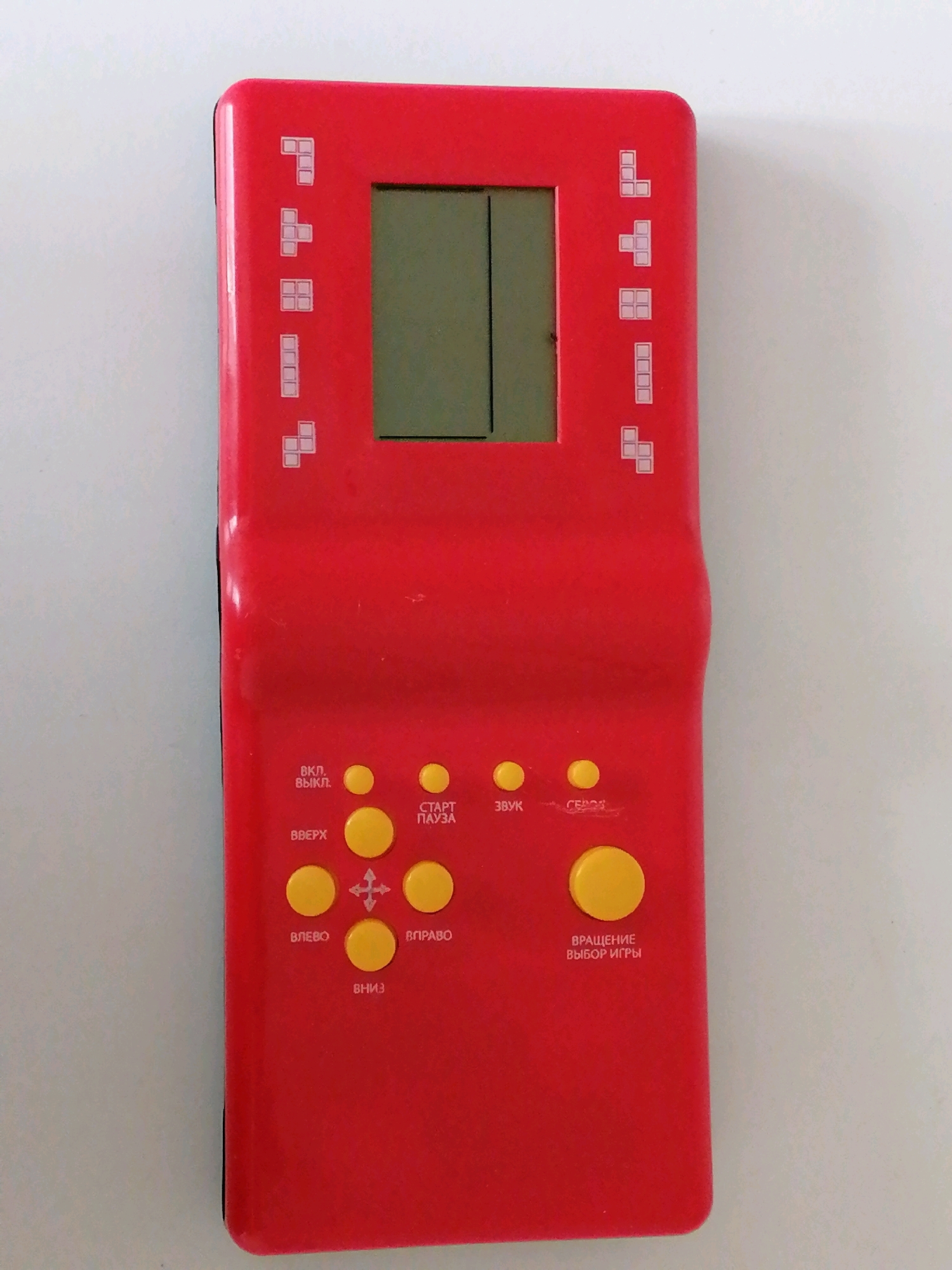 A red handheld electronic game console is photographed from a top-down perspective against a light gray background. The console is rectangular with rounded corners and a curved indentation on the right side. A small, dark screen is located at the top center of the console. Below the screen are various yellow buttons with Russian text. The buttons are arranged in rows and columns, with a directional pad in the center. There are also decorative pixelated Tetris-like blocks printed on the console on either side of the screen. The console appears to be the primary subject of the image. There are no people or other objects visible in the scene. The environment is a plain, indoor surface. The image is well-lit and in focus. The console's design and the text on the buttons suggest it is a Tetris-style game. It is not possible to determine the location of Kaliningrad, Russia from this image.