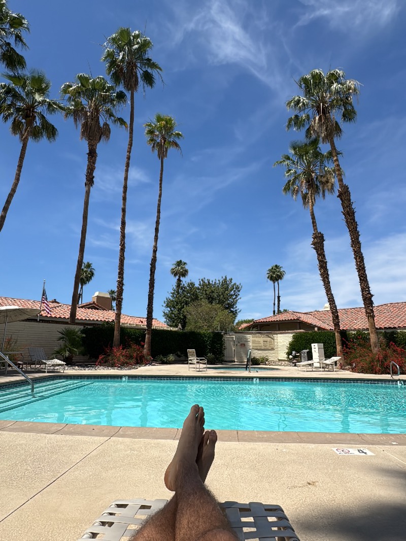 The image depicts a first-person perspective scene at a swimming pool in Palm Desert, United States. The viewer's legs and feet are visible in the foreground, resting on a white plastic lounge chair. The pool water is clear and turquoise. In the background, there are palm trees set against a clear blue sky with scattered clouds. There are low-lying buildings with red-tiled roofs behind the pool. An American flag is visible on one of the buildings. Landscaping including bushes, shrubs, and a few trees can also be seen in the background.