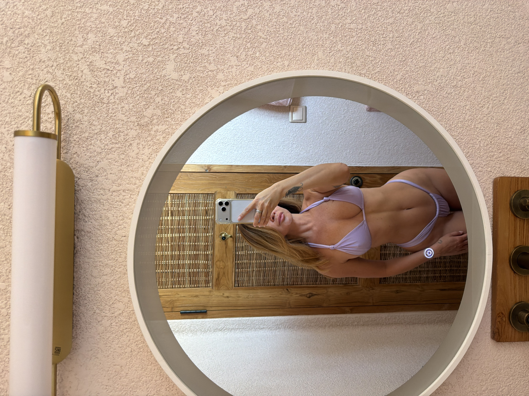 An indoor full-shot shows a woman taking a mirror selfie, reflected horizontally in a circular mirror against a textured wall.
The central subject is a woman, partially visible in the mirror, wearing a light purple string bikini. She has blonde or light brown hair and is holding a silver smartphone (likely an iPhone in a clear case) to take the photo. A ring is visible on her left-hand ring finger, and a tattoo, possibly a feather design, is on her right forearm. Her midriff and legs are visible, showing a fit physique.
The mirror itself has a light-colored, possibly off-white or beige, frame. Reflected behind the woman is a wooden cabinet or door with woven rattan panels, suggesting a natural or tropical aesthetic. A white wall and a light switch are also visible in the reflection.
Outside the mirror, the wall is textured and light-colored, possibly a pale pink or peach. To the left, a modern wall sconce with a curved gold/brass arm and a white cylindrical light fixture is mounted. The text "SNI" is visible on the base of this sconce. To the right, a wooden panel with brass-colored fixtures, possibly hooks or controls, is visible. A glowing white power symbol icon appears on the mirror surface near the woman's right arm, suggesting a touch control for a mirror light.
The scene conveys a casual moment, possibly within a resort or hotel room, consistent with the location context of Mengwi, Indonesia, given the natural materials like rattan and wood. The overall ambiance is bright and clean.
