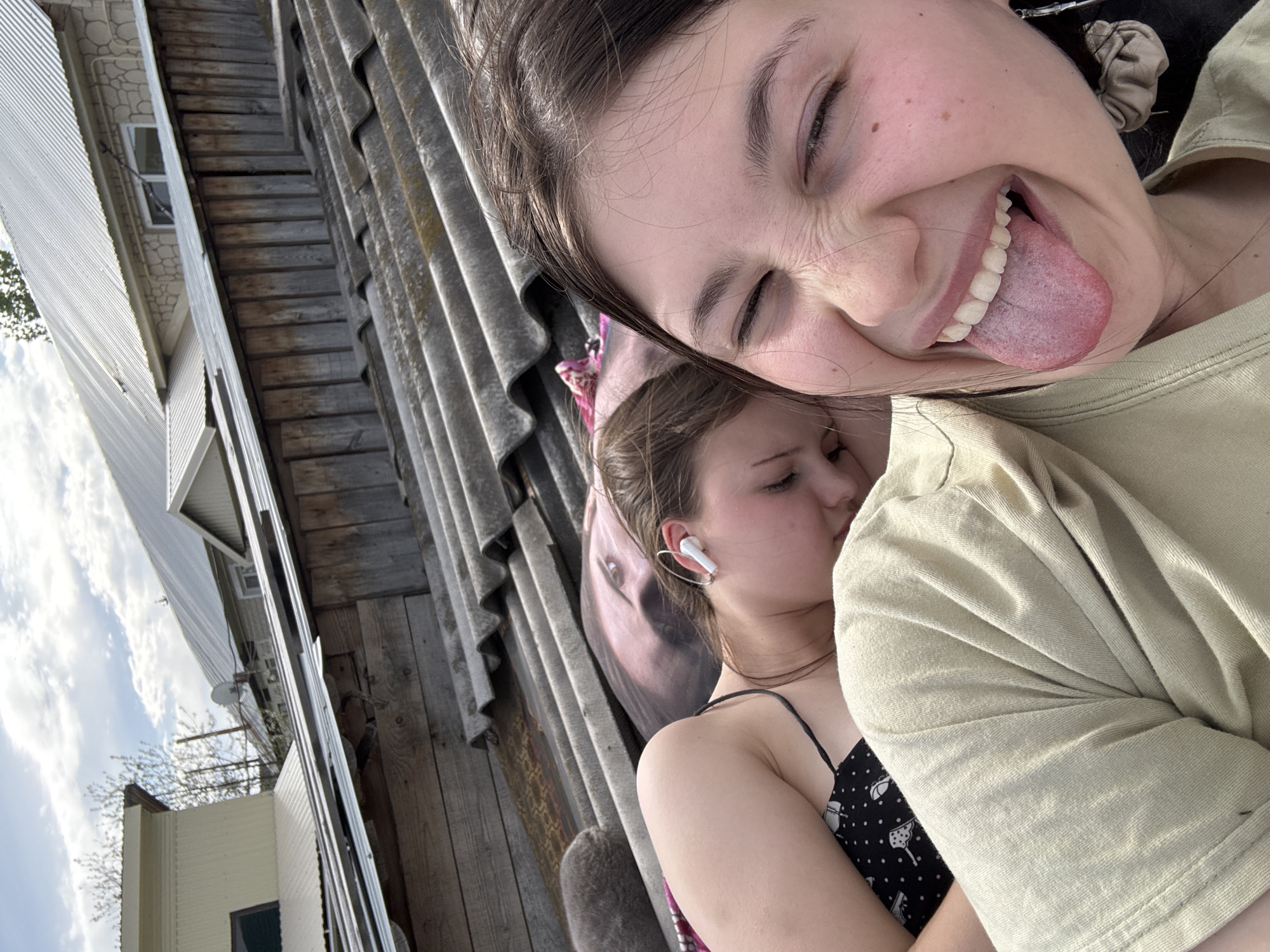 Two young women are in an embrace in an outdoor location. One woman is pulling a silly face and sticking out her tongue. The other woman appears to be sleeping or resting, and wearing an earbud. The background contains corrugated metal and wooden panels, as well as buildings, trees, and the sky. The location is Фоки, Russia.