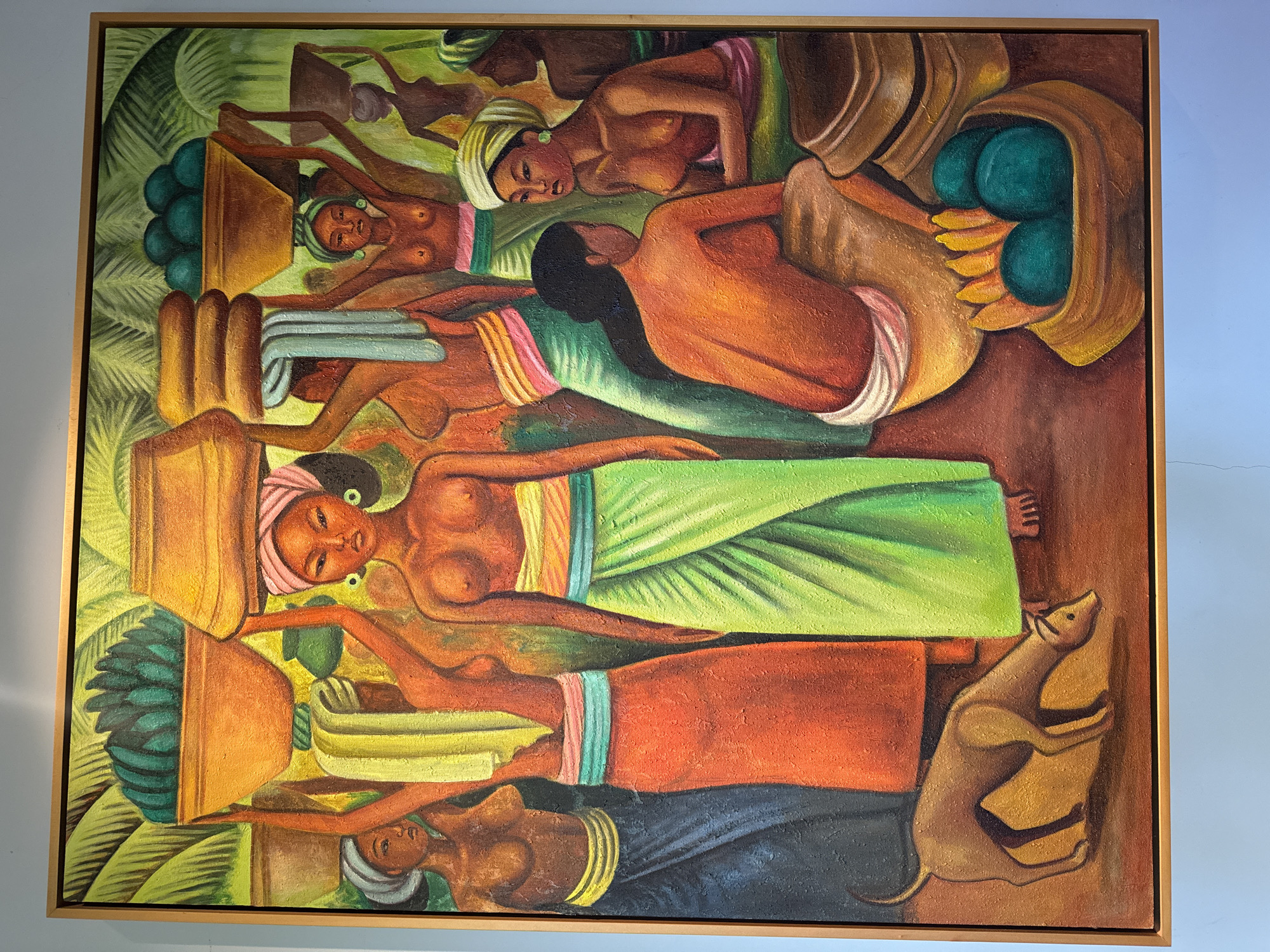 The image shows a painting in a wooden frame. The painting depicts a scene with multiple women, possibly Indonesian, carrying produce in containers on their heads. They wear turbans and brightly colored wraps. Some containers hold green fruits and bananas. A dog stands to the lower right. Green foliage is in the background. The artwork is associated with the Ubud District, Indonesia.