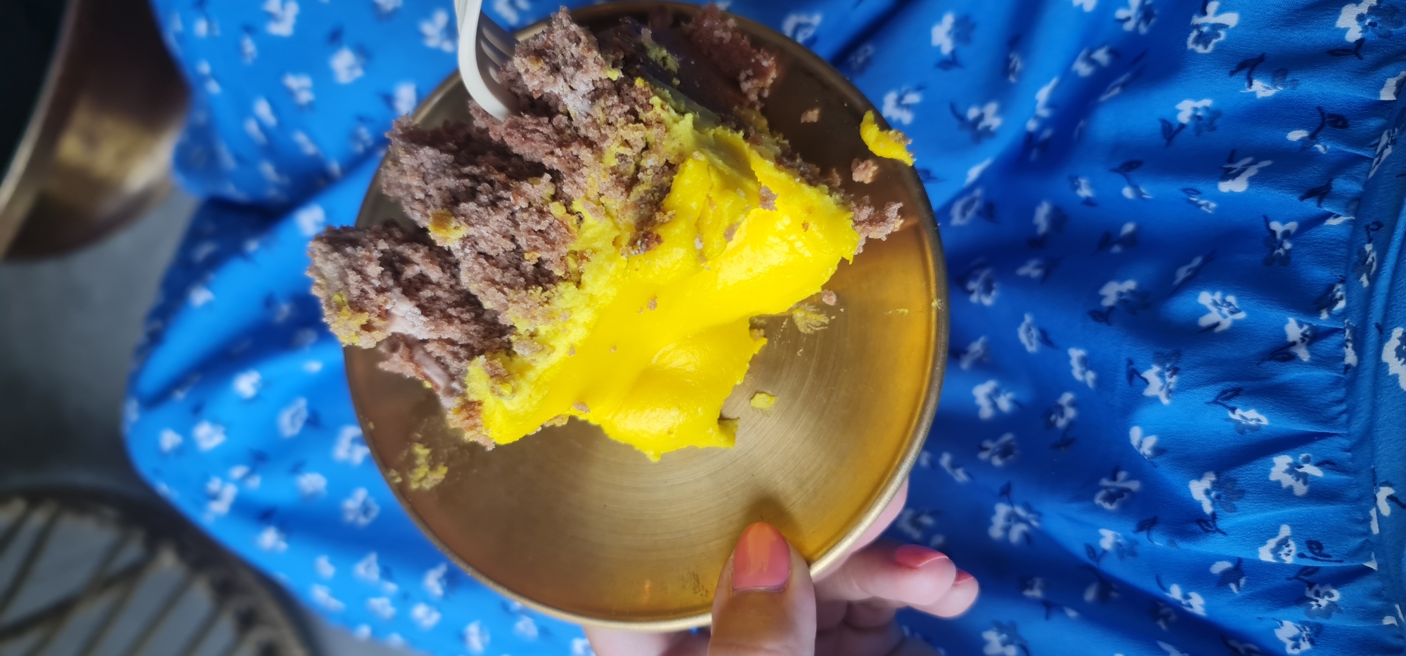 A high-angle shot shows a person holding a small gold plate with a piece of cake on it. The cake appears to be chocolate with a bright yellow layer, and a white plastic fork is visible. The person holding the plate has pink nail polish. The background is a vibrant blue floral print fabric, possibly a dress. The setting doesn't provide enough information to determine if the location is in Đà Nẵng, Vietnam. The composition focuses on the cake and plate.