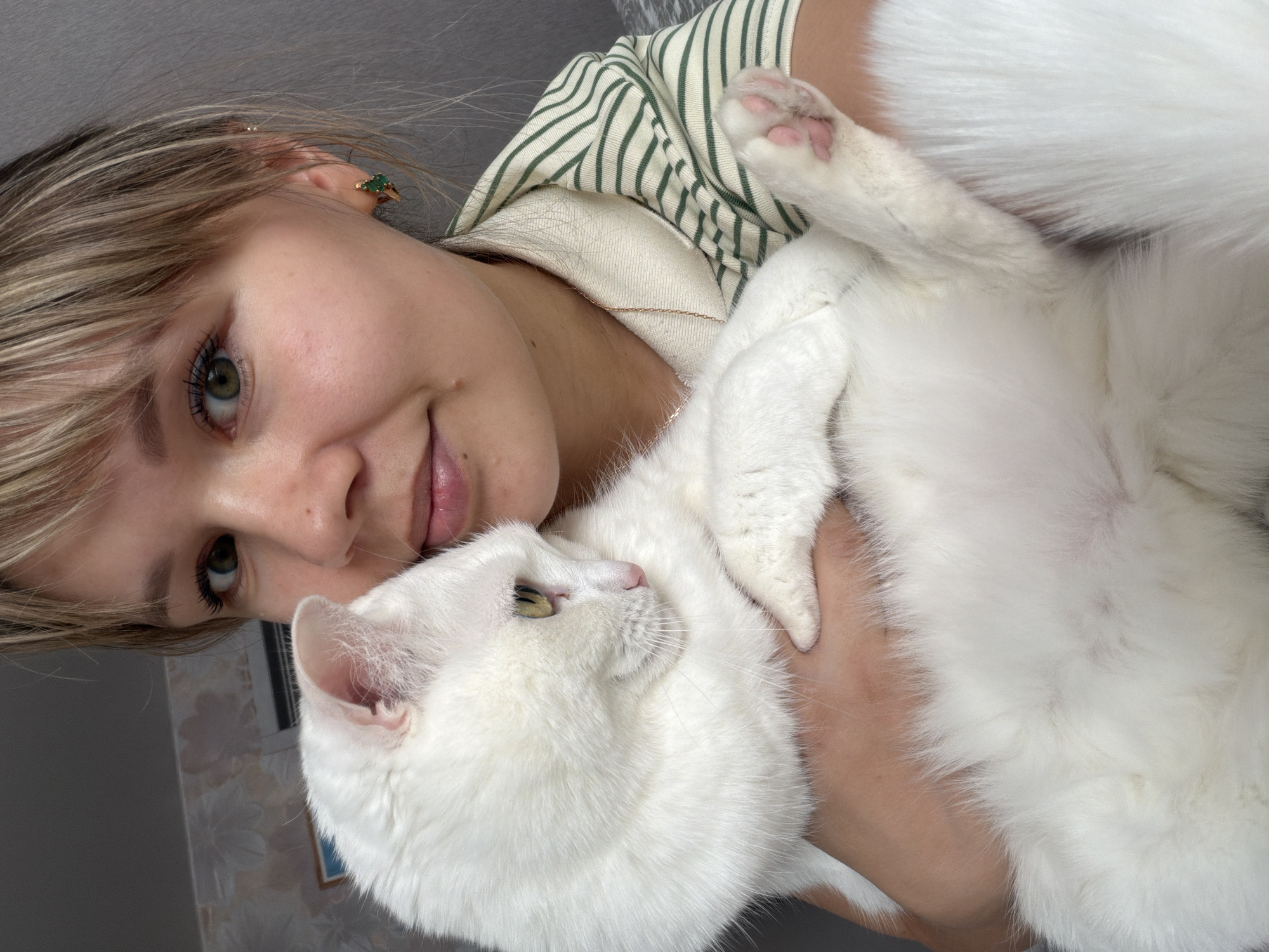 A close-up, tilted portrait captures a young woman cuddling a fluffy white cat indoors. The woman, with blonde hair and green eyes, looks towards the camera with a gentle, faint smile. She is wearing a cream-colored top with green vertical stripes and a delicate gold-toned necklace. A small earring with green stones is visible in her right ear.
She is embracing the white cat, which has striking yellow-green eyes and a pink nose. The cat appears relaxed and comfortable, looking slightly upwards and to the side, with one paw extended, showing its pink paw pads. The soft fur of the cat takes up a significant portion of the right side of the image.
The background is simple, featuring a plain light-colored wall on the upper left and a wall with a subtle patterned wallpaper on the lower right, suggesting a home environment. The lighting is bright and even, indicating it is likely daytime. The overall impression is one of warmth, affection, and companionship between the woman and her pet. There is no visible text in the image.