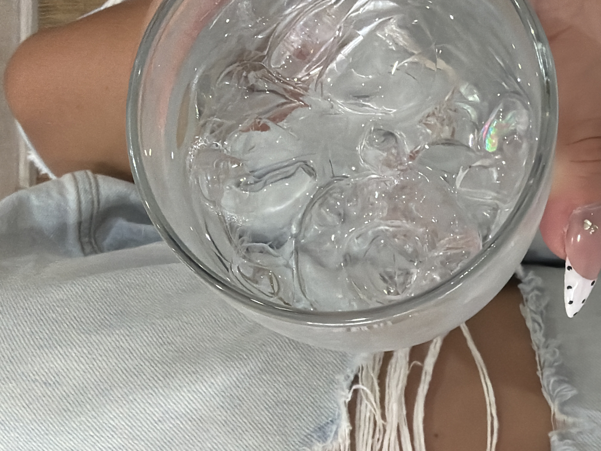 An overhead shot depicts a glass filled with ice water, held in a person's hand, with the backdrop of distressed light-wash denim and fringes. The person's fingernail is visible, adorned with a French tip design, which is primarily white with black polka dots near the tip and a beige base, and there are at least two small gems near the cuticle. The scene is likely indoors. There are no definitive clues to place this image in North Kuta, Indonesia.