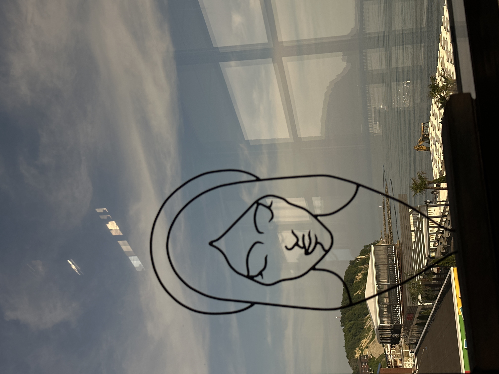 This image depicts a coastal scene in Lermontovo, Russia, viewed through a window adorned with a linear artwork. In the immediate foreground, a black outline drawing of a woman's face with closed eyes, long hair, and a circular element resembling a halo is present on the windowpane.
The upper portion of the image shows a cloudy sky, transitioning from light blue to a pale greyish-white, with visible stratocumulus cloud formations. Several distinct, rectangular, and illuminated reflections are visible against the sky in the upper left and center, suggesting internal light sources or reflections from an adjacent structure.
Outside the window, a prominent, tree-covered hill or cliff rises on the left side, supporting a small white structure at its summit. Below the cliff, a complex of buildings lines the shoreline. A large, rectangular structure with a white roof and dark, enclosed sides is elevated on stilts, extending over or very close to the water. Below this, multiple levels of white-canopied or covered outdoor areas with seating are visible, interspersed with several palm trees. A yellow excavator is positioned on the sandy or stony shoreline in front of these structures.
The majority of the right half of the image features the calm, grey-blue water of the sea. A long, narrow rock breakwater or pier extends into the sea from the mid-right, culminating in a small, light-colored beacon or navigational aid. Reflections of window frames from the interior create a distinct grid pattern over the sea and the hazy outlines of distant mountains or landmasses across the water. In the bottom left corner, a dark, flat rooftop with a visible green, yellow, and orange striped edge is partially captured. No human figures are clearly identifiable in the scene.