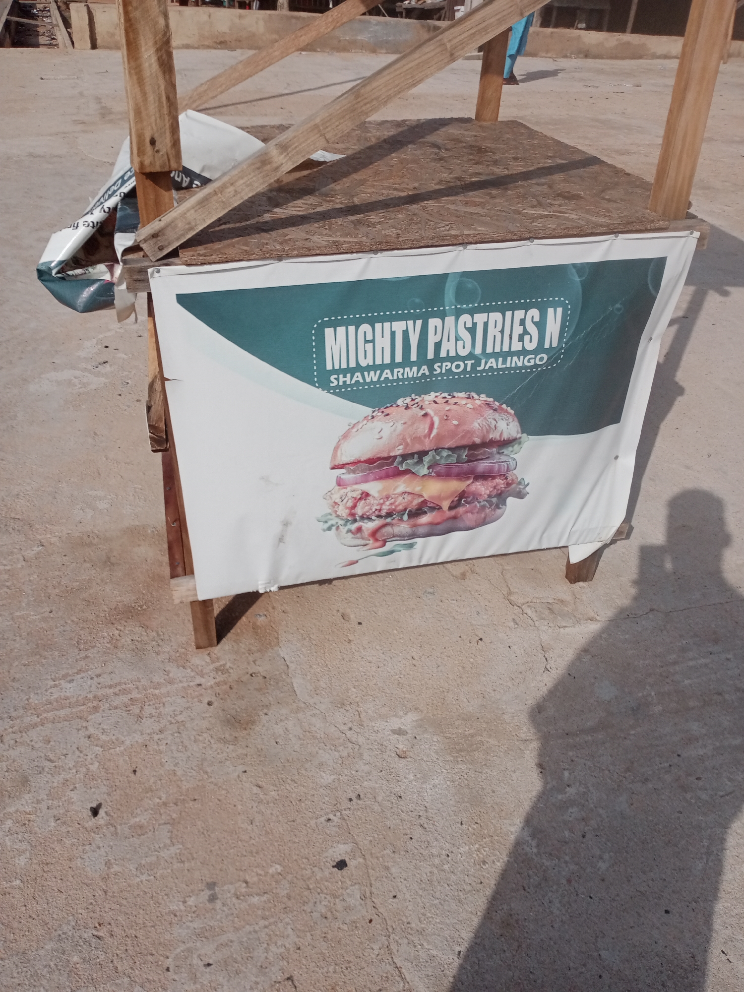 A close-up, eye-level shot captures a makeshift food stall in Jalingo, Nigeria. The stall features a wooden frame supporting a plywood countertop. Hanging from the front of the stall is a large banner advertising "MIGHTY PASTRIES N SHARMA SPOT JALINGO." The banner prominently displays a mouth-watering image of a large burger. The stall is situated on a rough, concrete surface. To the right of the stall, a person's shadow is cast onto the ground, suggesting it is a sunny day. In the background, out of focus, there are structures that appear to be part of a market or a similar outdoor commercial area. The overall impression is one of a humble, yet inviting, street food vendor.