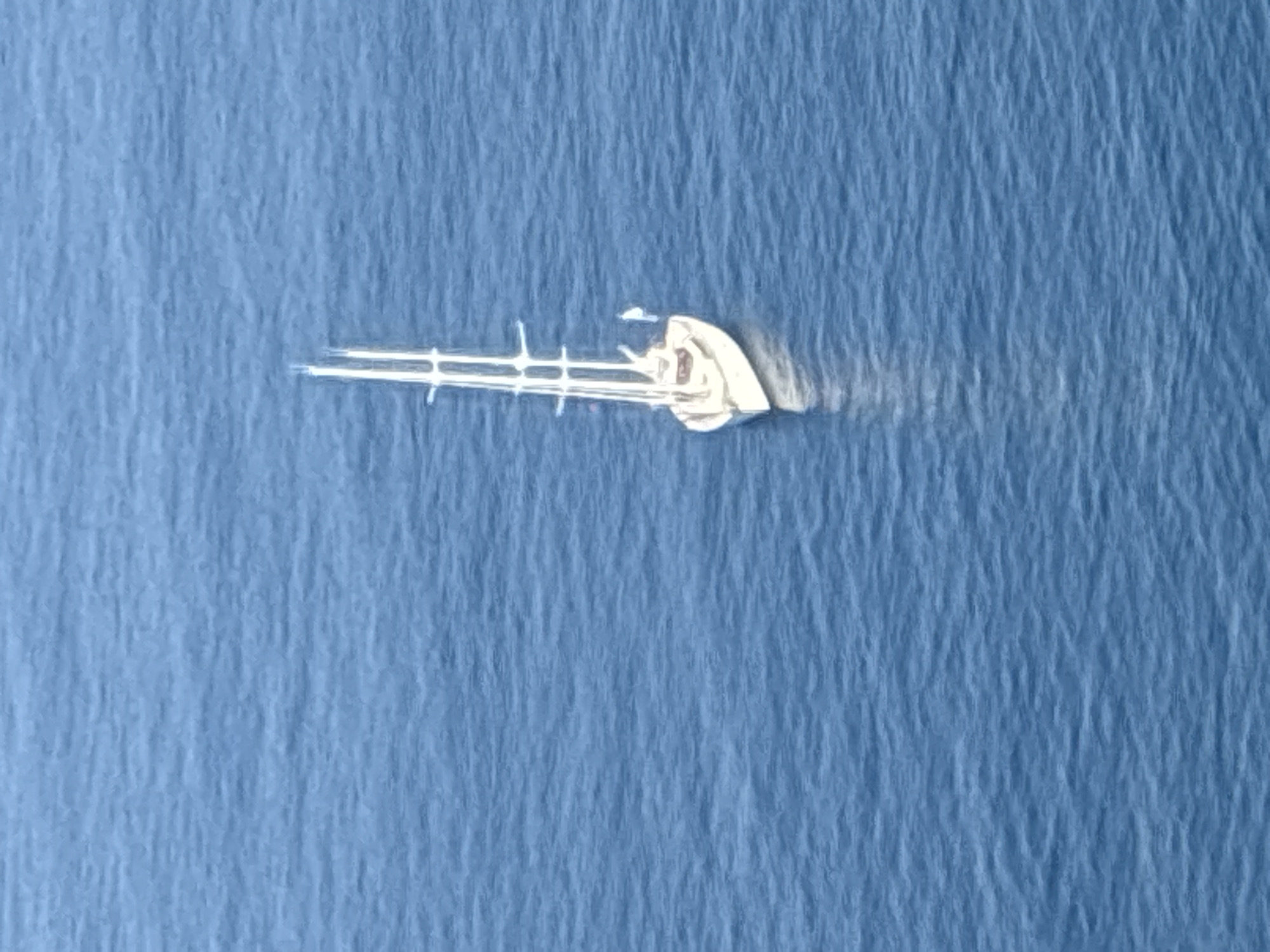 An aerial, high-angle shot shows a white sailboat with two masts floating on a vast expanse of blue ocean. The boat is positioned in the center-right of the frame. The water is calm with subtle ripples, creating a textured surface. The sky is not visible. The scene is tranquil and devoid of any human activity or other vessels. The location is unspecified.