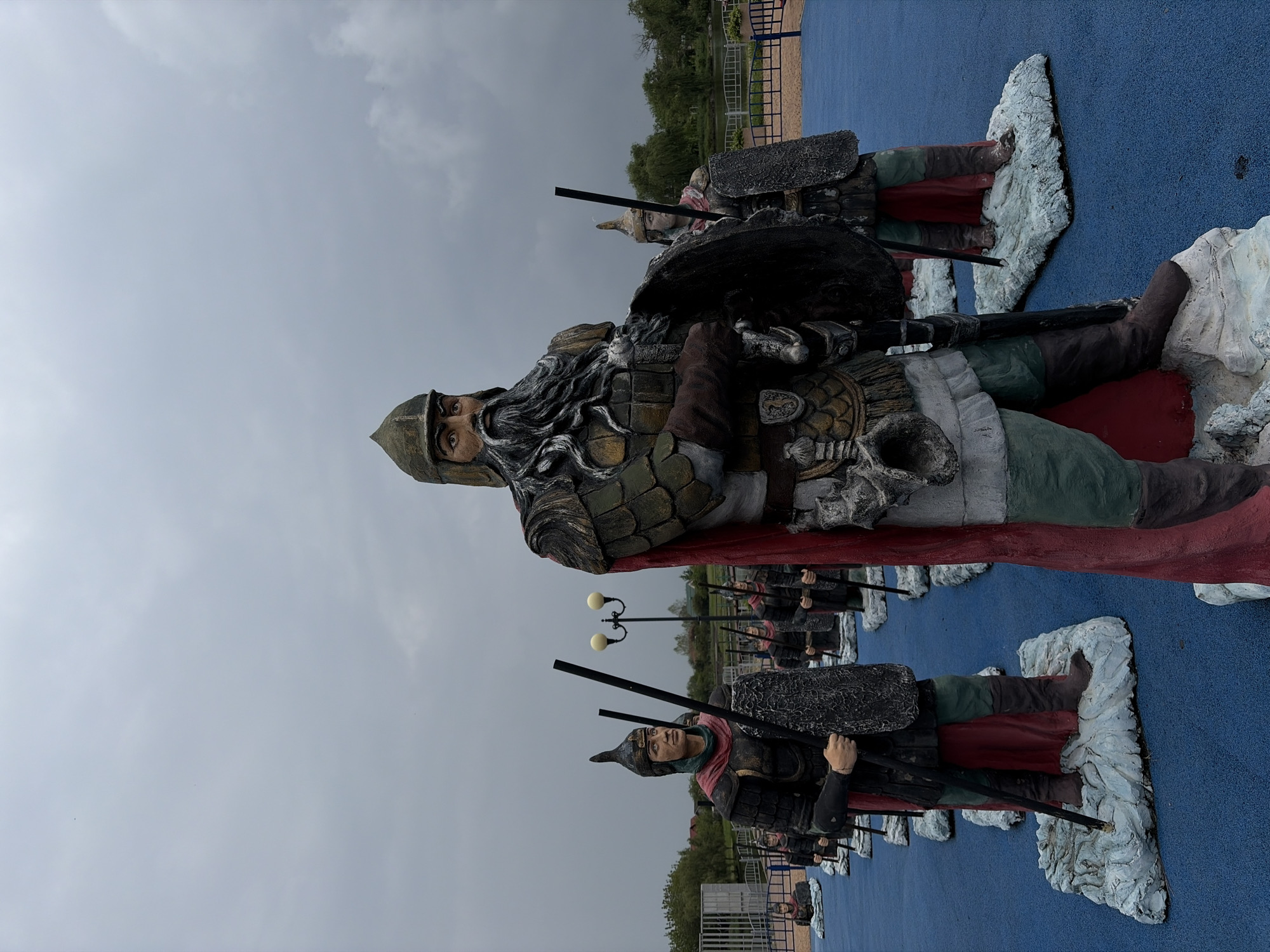 The image shows a row of painted warrior statues, possibly made of concrete or a similar material, standing on a blue surface. The statues appear to be of medieval or ancient warriors, with helmets, armor, shields, and weapons. The sky is overcast, suggesting a cloudy day. The location appears to be a park or outdoor exhibition in the city of Unknown, Unknown. There are street lamps and trees in the background.