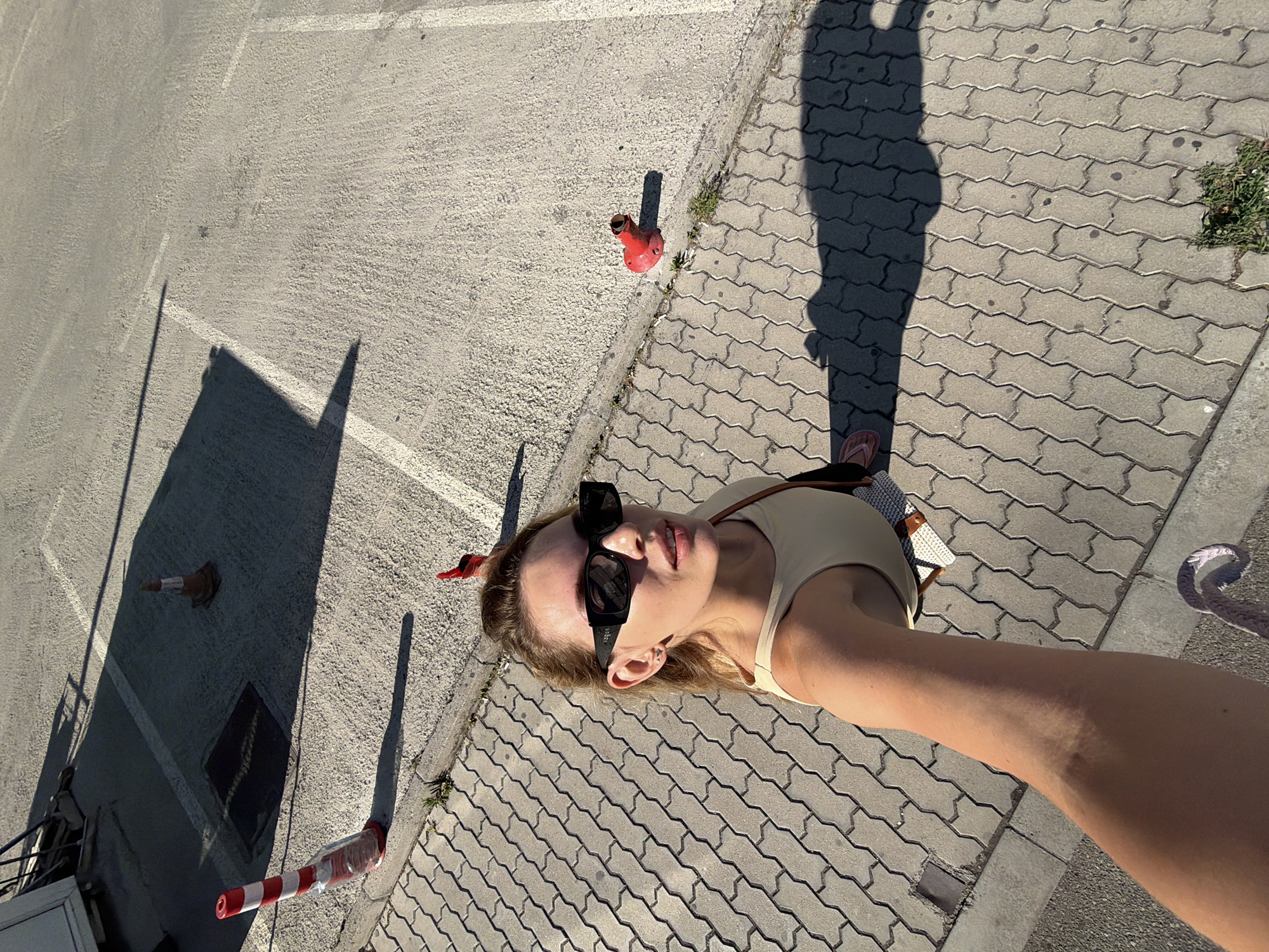 A high-angle, eye-level selfie captures a woman wearing black sunglasses, a beige tank top, and a cross-body bag standing on a paved area in Budva, Montenegro. The ground is a mix of rough concrete and interlocking brick pavers. In the background, orange traffic cones are visible, with one particularly prominent striped cone near the left edge of the frame. A red object, possibly a decorative bollard, sits on the concrete to the right. Shadows from the woman and other unseen objects are cast across the paved surface, indicating a sunny day. The overall scene suggests an outdoor urban or semi-urban environment.