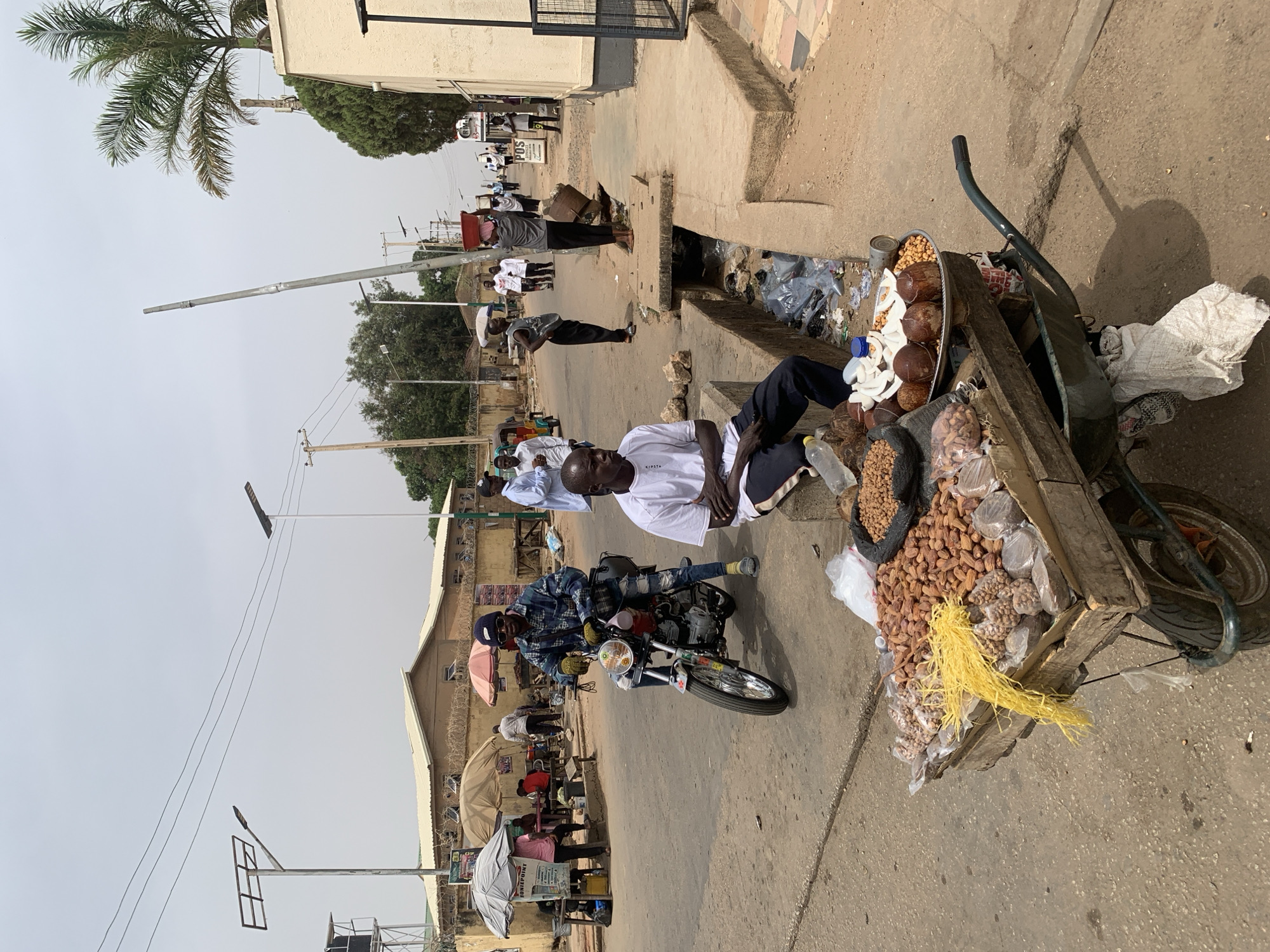 This image captures a bustling street scene in Jos, Nigeria, likely during the day. The primary focus is a street vendor sitting on the ground next to a wooden pushcart laden with various food items for sale.
**Subjects:** In the foreground, a man wearing a white shirt with "KIPSTA" visible on it and dark trousers sits beside a well-stocked pushcart. The cart holds whole and sliced coconuts on a metal tray, a large pile of peanuts, dried dates, and several clear plastic bags containing other snacks or nuts. A yellow, stringy material is also draped over the cart. In the mid-ground, another man on a motorcycle, dressed in a plaid shirt, jeans, and a dark beanie, is visible. Several other individuals are scattered along the street, some walking, others standing or gathered further down. A group of men in white robes or shirts can be seen in the distance. The scene features buildings with simple architecture, some with corrugated iron roofs, and utility poles with overhead wires. A palm tree is visible on the left, and other trees dot the landscape. A drainage ditch filled with trash runs alongside the road.
**Activity:** The central activity revolves around street vending, with the cart displaying goods for sale. People are engaged in daily commutes and activities on the street, with a motorcyclist present and others walking.
**Notable Details:** The time of day appears to be sunny and bright, suggesting mid-morning or afternoon with clear skies. The lighting casts clear shadows. The street is paved, with concrete sidewalks and a visible drainage system. The overall atmosphere is that of a typical urban or semi-urban street market.
**Visible Text:** "KIPSTA" is clearly visible on the sitting vendor's shirt. Further down the street, a sign with "POS" and some smaller, illegible text can be discerned. An umbrella in the background appears to have "UNILPINT" or similar text.