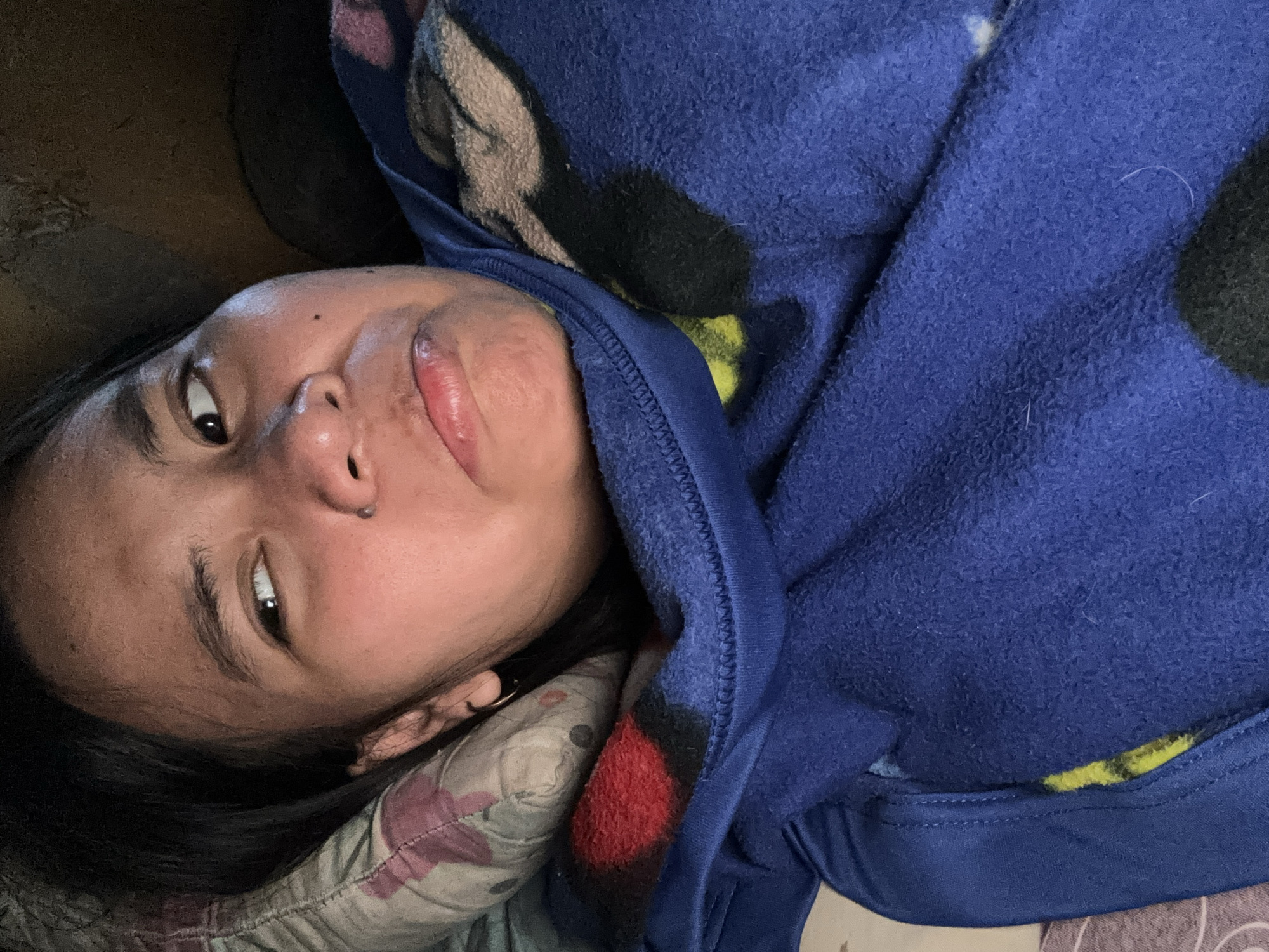 A close-up, overhead shot shows a person lying in bed, their face turned to the side with a slight frown. They are wearing a blue fleece hoodie with a cartoon character print. The background is dark and appears to be a wall. The image was taken indoors, in a bedroom located in East Jakarta, Indonesia.