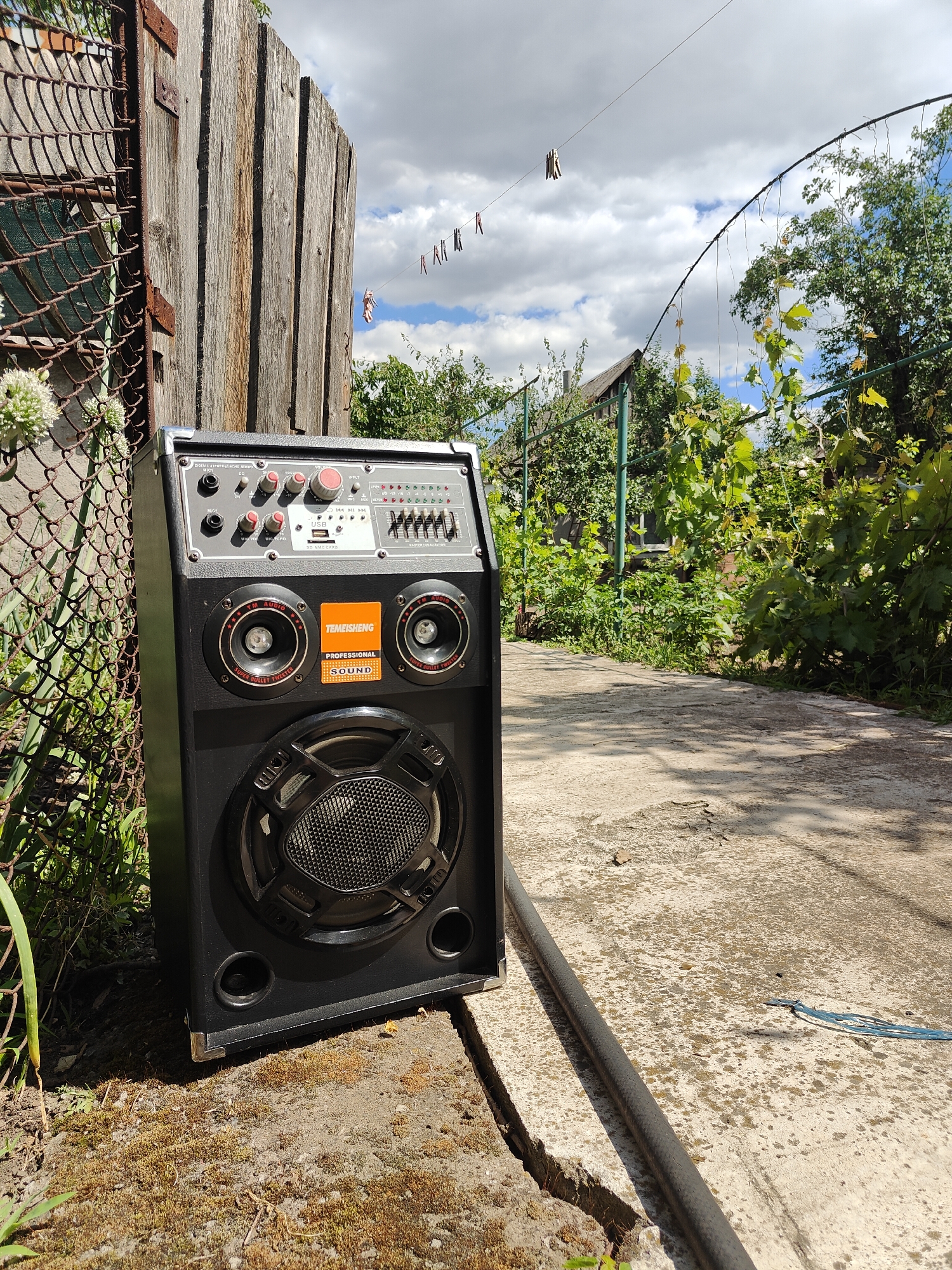 An outdoor scene in Kryvyi Rih, Ukraine, features a large black portable speaker situated on a concrete surface next to a weathered wooden fence and chain-link fencing. The speaker has multiple control knobs and buttons on its top panel, two tweeters, and a prominent central woofer. A garden path, overgrown with vegetation, extends behind the speaker. Clothes hang on a clothesline strung between two poles in the background, and trees and a house are visible beyond the garden. The sky is partly cloudy.