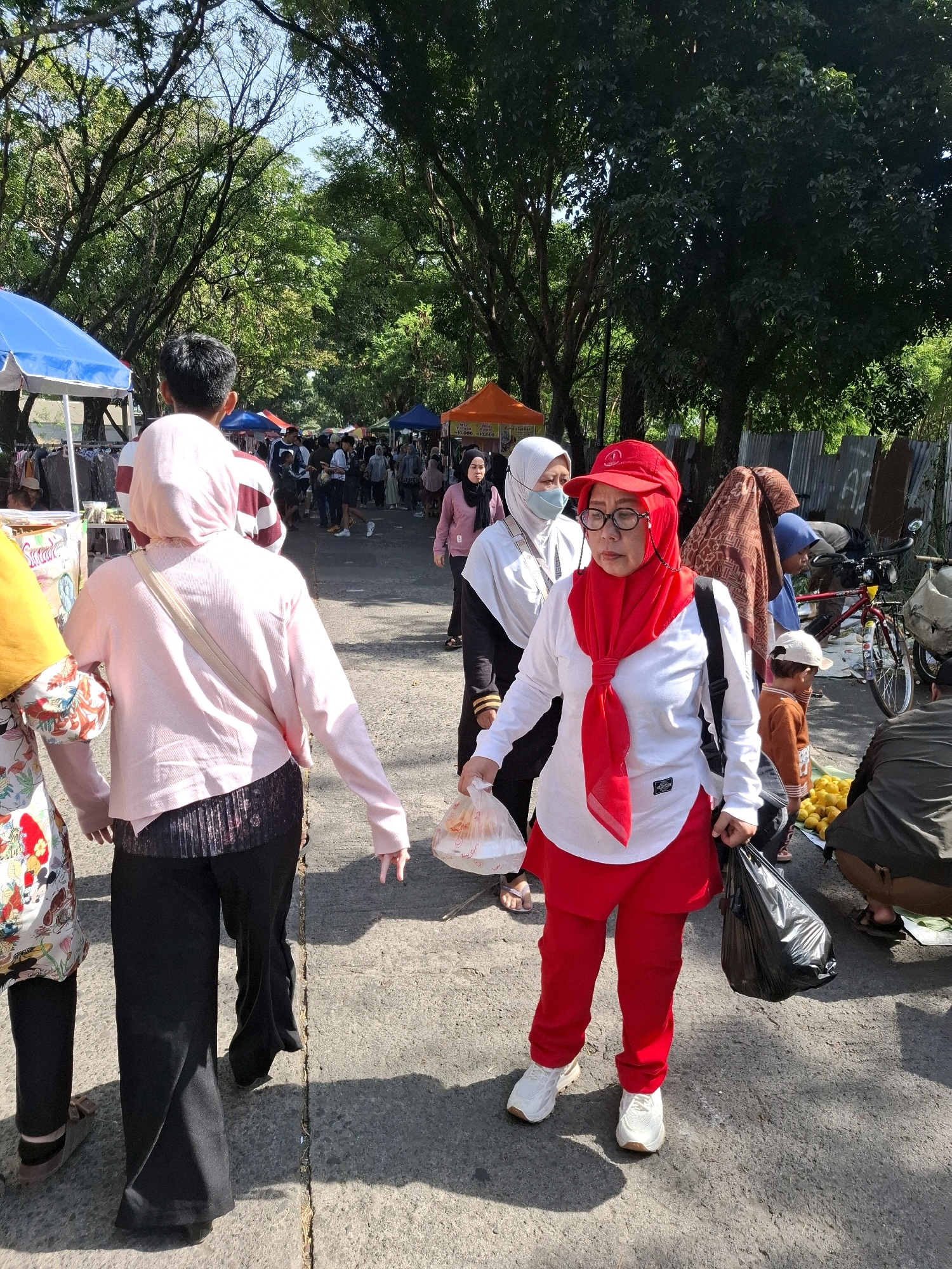 The image depicts a street market scene in Karangtengah, Indonesia, under a canopy of trees. In the foreground, a woman wearing a red hijab, red cap, glasses, and a red scarf over a white long-sleeved shirt and red pants is walking towards the camera, holding a plastic bag in her right hand and a black plastic bag in her left. To her left, another woman in a pink hijab and a light pink long-sleeved shirt with black patterned pants is walking away from the camera. To the right of the woman in red, a child wearing an orange shirt and a white cap is standing near a pile of yellow fruits, possibly lemons. Behind them, several other individuals are visible, some walking and others browsing at market stalls. The market appears to be set up along a paved road, with tents and structures lining the sides. A bicycle with a basket is parked on the right side of the image. The overall atmosphere suggests a busy, outdoor market day.