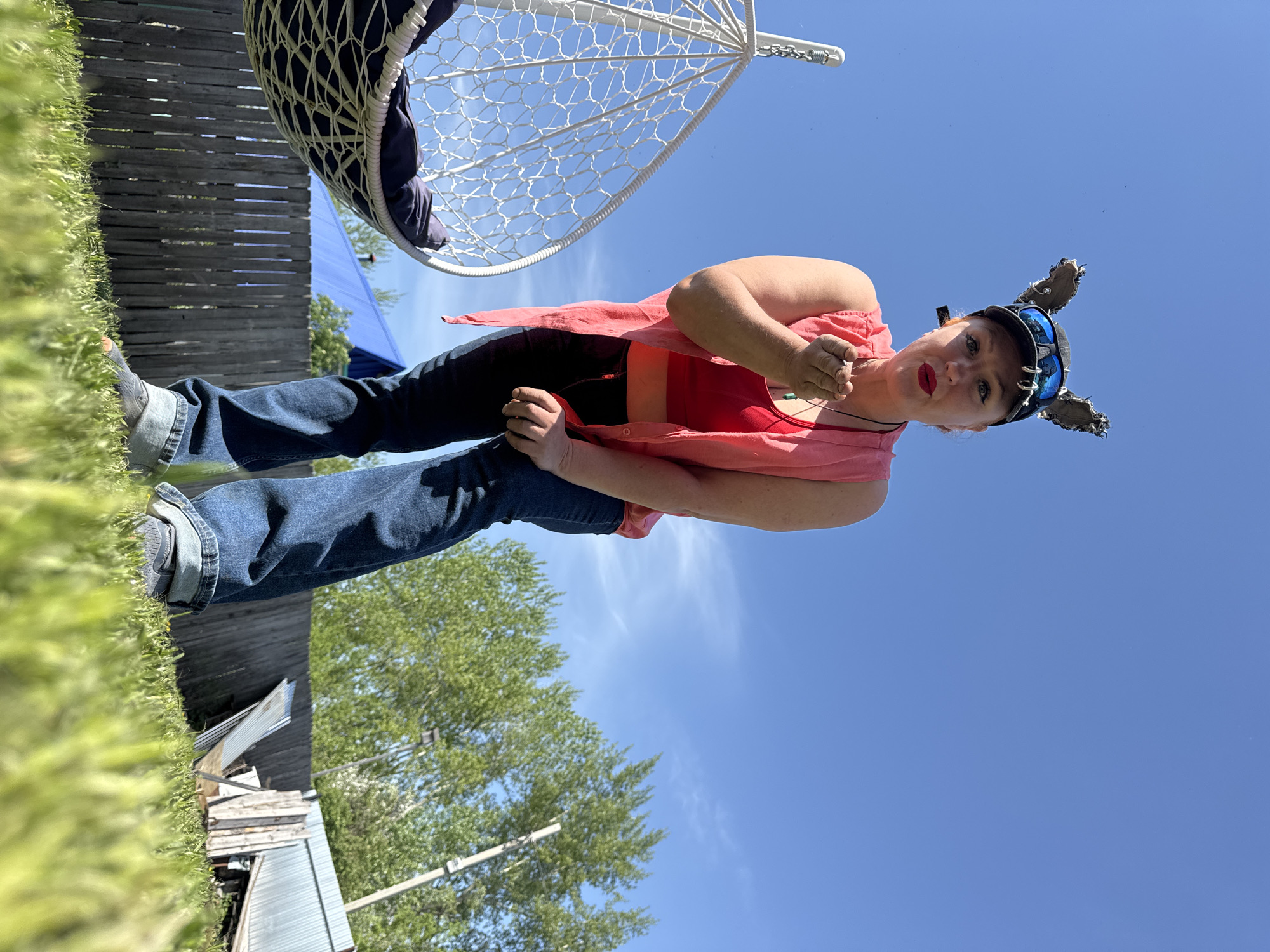 The image shows a woman standing outdoors on a sunny day. She is wearing jeans, a red top, a pink open shirt, and a cap with decorative ears. She has red lipstick on and is holding something small in her hand, near her mouth. Behind her is a blue sky with some wispy clouds. To her left is a white hanging chair. The environment includes a wooden fence and some vegetation, possibly grass and trees. In the background, there is a blue structure and a dilapidated shed-like building. The location appears to be in a yard or garden setting. The city, Chaikovsky, Russia, is a possible, but not provable, location.