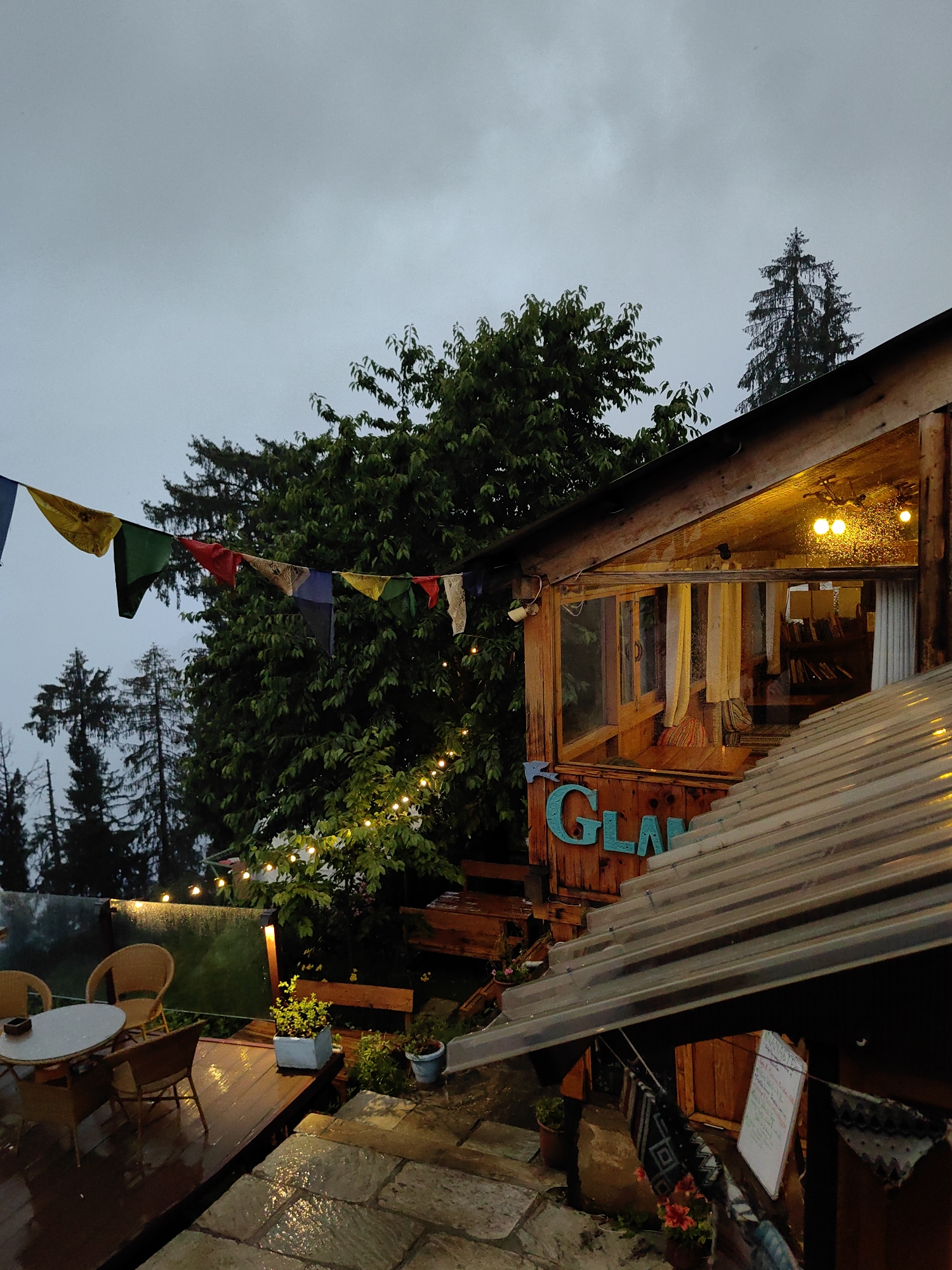 The image depicts an outdoor seating area and the exterior of a wooden structure, likely a cafe or guesthouse, in Siumsa, India, under an overcast sky. Prayer flags in various colors, including yellow, green, red, blue, and white, are strung across the scene. String lights are visible, illuminating trees and the building. The wooden structure has large windows, some with yellow curtains, and a sign with teal-colored lettering that appears to read "GLAM." Inside the building, bookshelves are visible through the windows.
In the foreground, there is a wet wooden deck with a small round table and two wicker chairs. Adjacent to the deck is a stone patio, also wet, suggesting recent rainfall. Several potted plants are scattered around the area, including a small green plant in a grey pot and a red flowering plant in a brown pot. The ground is visibly wet and reflective. The overall atmosphere is muted, possibly due to the cloudy weather. No people are visible in the image. The location appears to be a scenic spot, possibly in a mountainous or hilly region given the presence of tall evergreen trees in the background.
