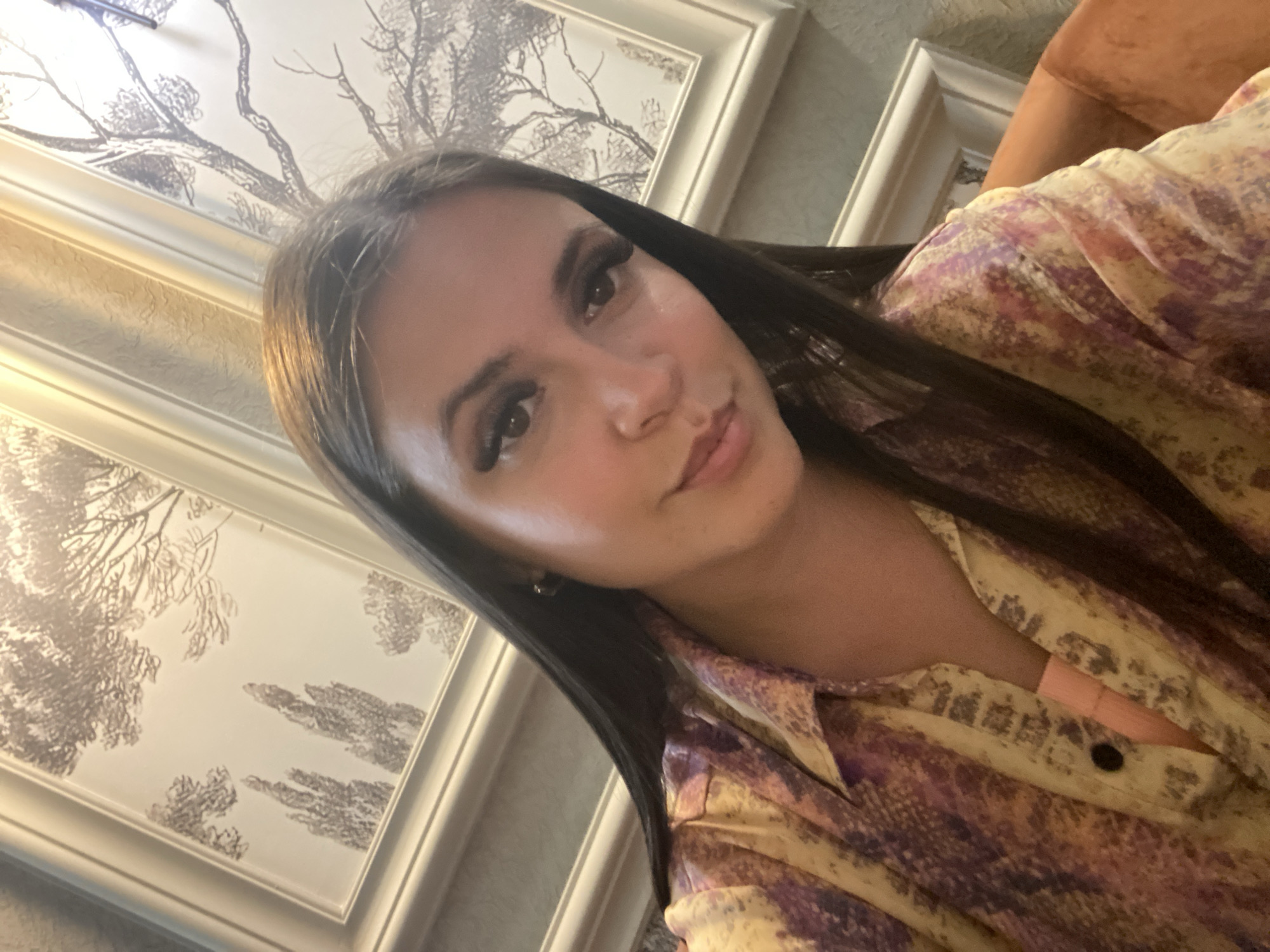 The image shows a close-up, selfie-style shot of a young woman with long, dark hair, wearing a patterned shirt. She is looking towards the camera with a slight smile. Behind her, there are framed pictures on the wall with a tree motif. The lighting suggests an indoor setting, possibly a restaurant or cafe. There is no discernible information in the image that allows for identifying its location in Makhachkala, Russia, or any specific details about the surrounding city.