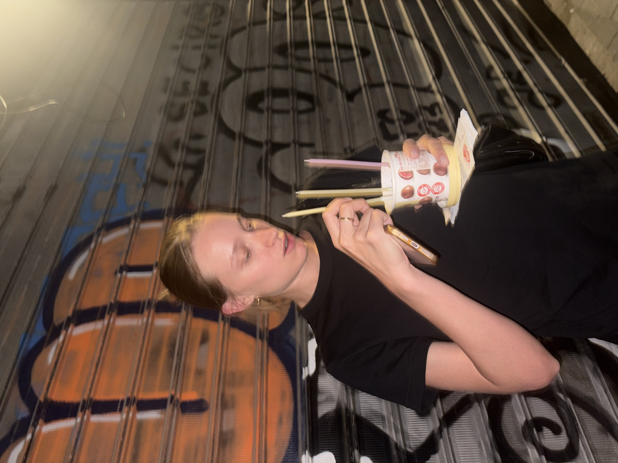 A young woman with blonde hair pulled back stands in front of a metal security gate covered in graffiti. She wears a black t-shirt and is holding a white paper cup with several straws and a small paper holder underneath. She is also holding a smartphone in her right hand. The graffiti on the gate is colorful, with large orange and blue spray-painted shapes on the left, and black and white graffiti on the right. The lighting suggests it is nighttime, with a bright light source from the upper right. The scene appears to be in an urban outdoor environment in Tel-Aviv, Israel, based on the characteristics of the security gate and graffiti common to the city.
