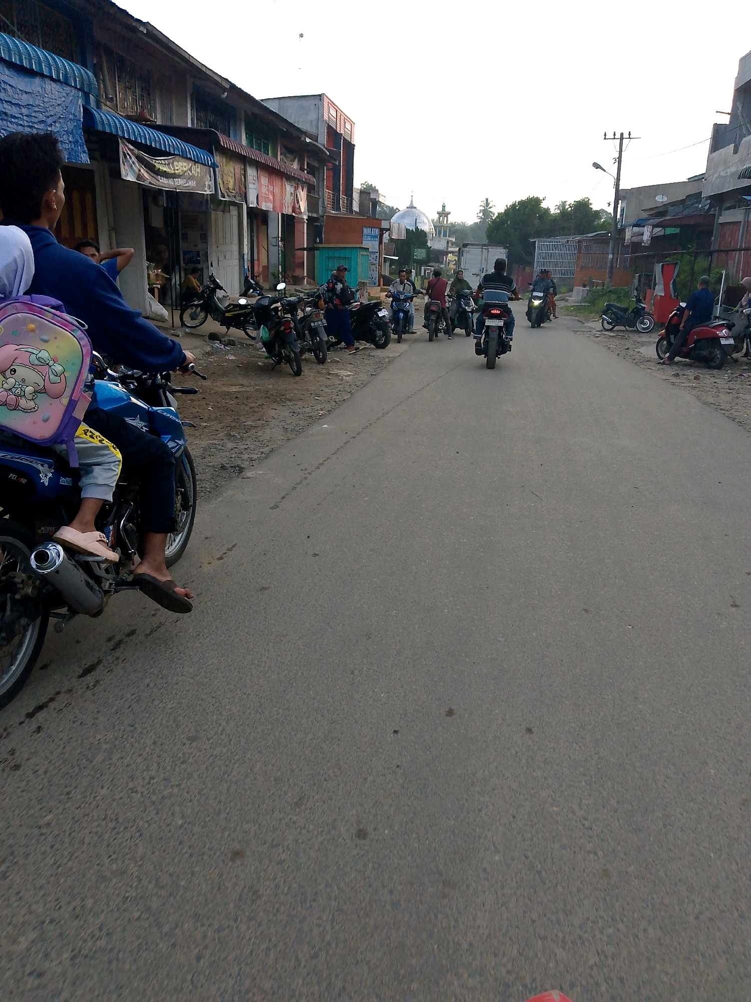 The image depicts a street scene in Dolok Merawan, Indonesia. In the foreground, a person on a blue motorcycle is riding down a paved road. They are wearing dark pants and a blue jacket, with a backpack adorned with a cartoon character on their back. To the left of the motorcycle, a row of buildings with awnings lines the street, suggesting a commercial area. Parked on the side of the road are several other motorcycles, some occupied by people. Further down the road, more motorcycles are in motion, with riders heading in the same direction. The background shows more buildings, trees, and utility poles. The sky is overcast, indicating a potentially cloudy or hazy day. The overall impression is one of daily life and transportation in a town in Indonesia, with a strong emphasis on motorcycle use.