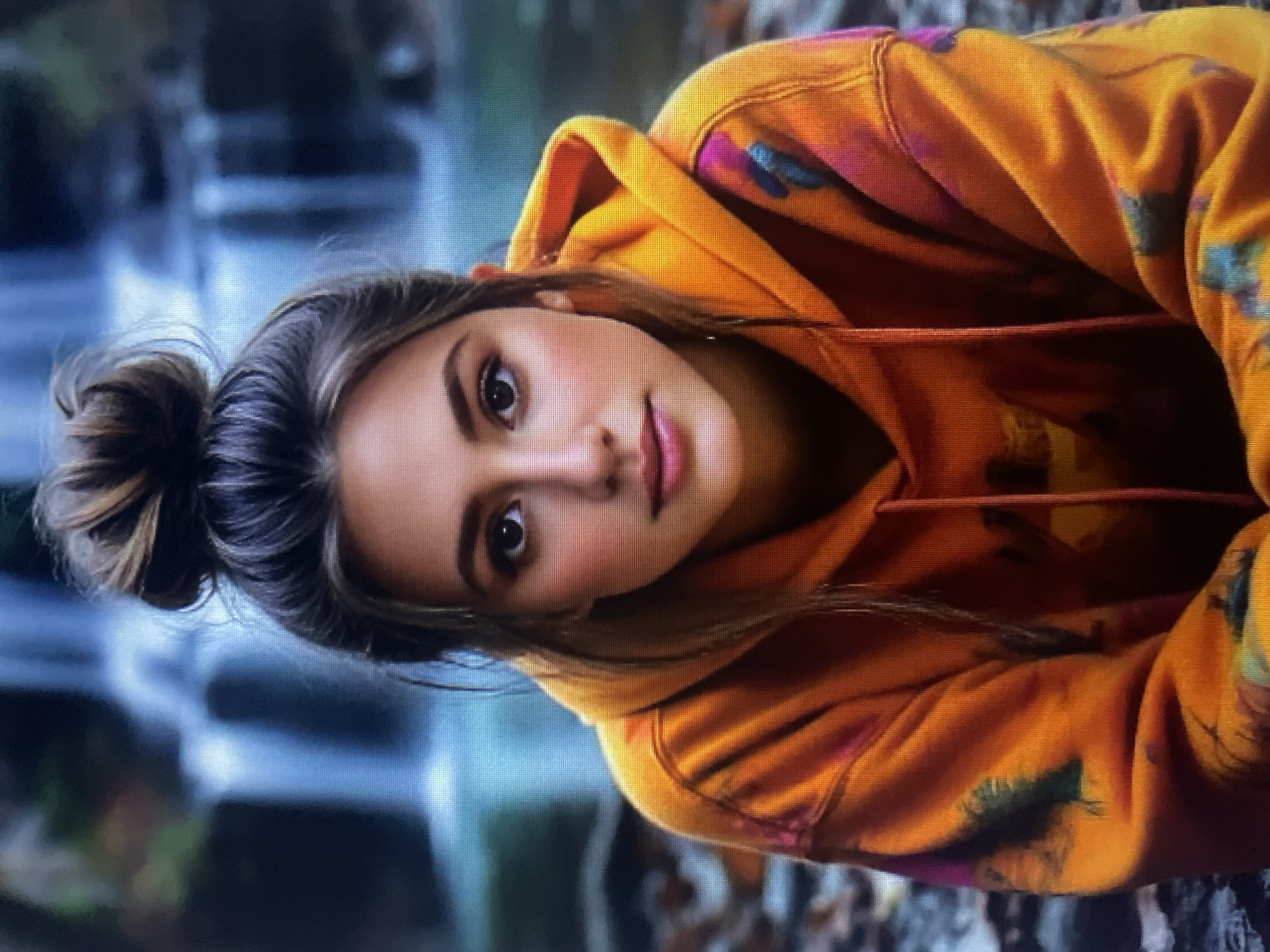 A portrait of a young woman with her hair in a messy bun. She is wearing an orange tie-dye hoodie. The background appears to be a natural setting with a waterfall. The image does not provide any information about the city of Kołobrzeg, Poland, nor any specific location within it.