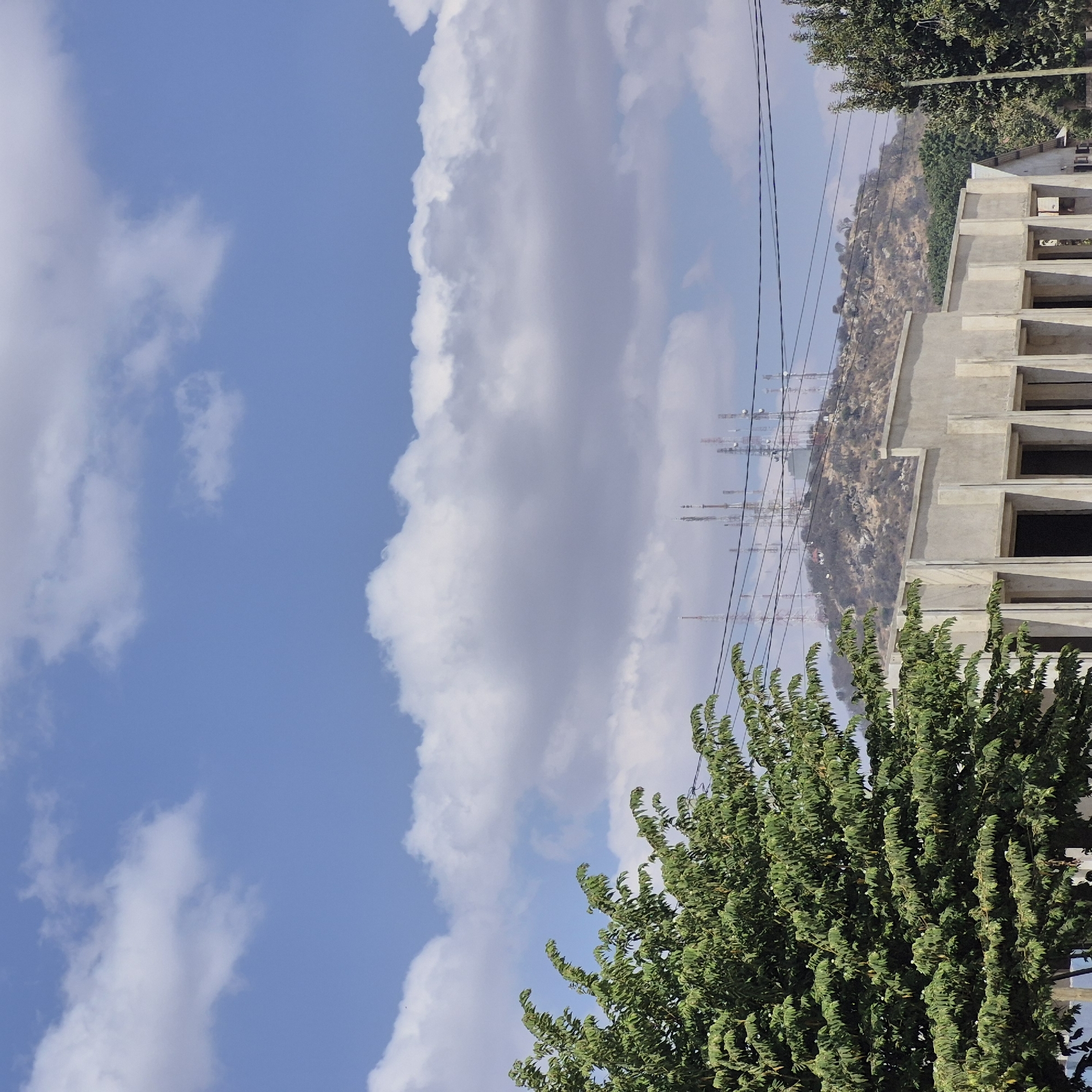 This image depicts an outdoor scene under a bright blue sky with scattered white clouds, likely in Dodoma, Tanzania. In the foreground, the upper branches of a lush green tree with dense foliage are visible on the left. A modern, unfinished concrete building with exposed structural elements and dark openings for windows or doorways occupies the lower central and right portions of the frame. Beyond the building, a dry, rocky hillside rises, dotted with sparse vegetation. The crest of the hill is adorned with a cluster of tall telecommunications towers, some of which have visible antennas and supporting structures. Power lines are strung across the scene, traversing the middle ground between the hill and the foreground. The overall environment suggests a sunny day in a semi-arid urban or peri-urban setting. There are no people visible in the image.