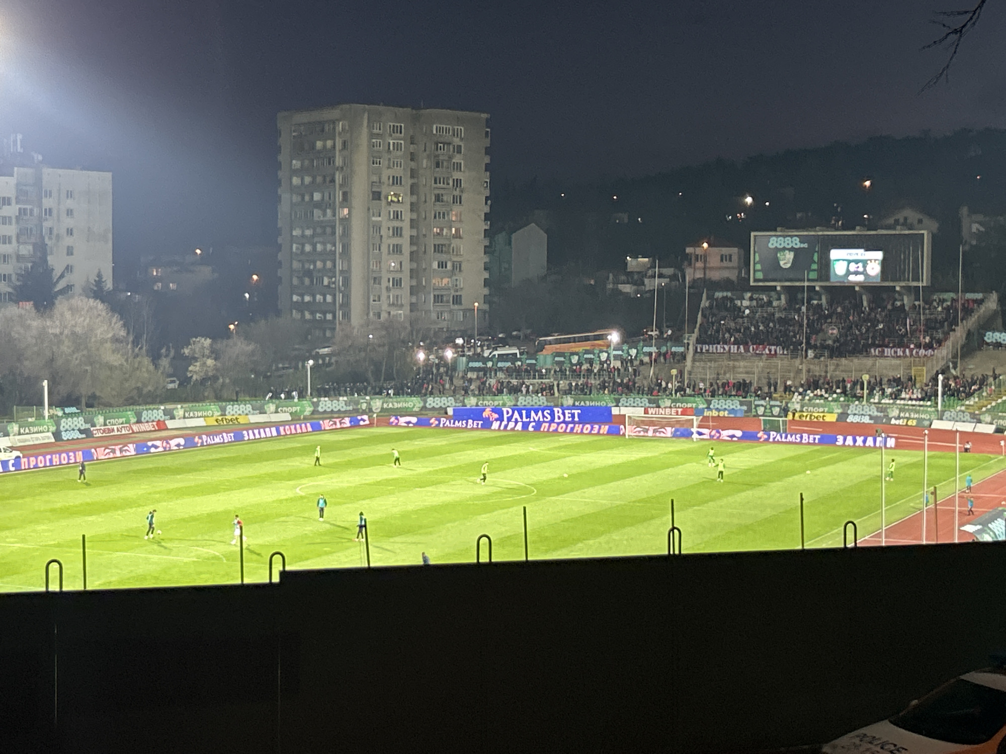The image shows a nighttime soccer game in progress at a stadium. The stadium is illuminated by floodlights, and a large crowd is visible in the stands. Advertisements line the field perimeter, primarily for "PALMS BET" and "8888." A large scoreboard displays the game information. In the background, several apartment buildings rise into the night sky. At the bottom of the frame, a dark wall is visible, and the corner of a police car is visible in the bottom right. The location appears to be in Stara Zagora, Bulgaria, based on stadium advertisements. There are players on the field, and the game is active.