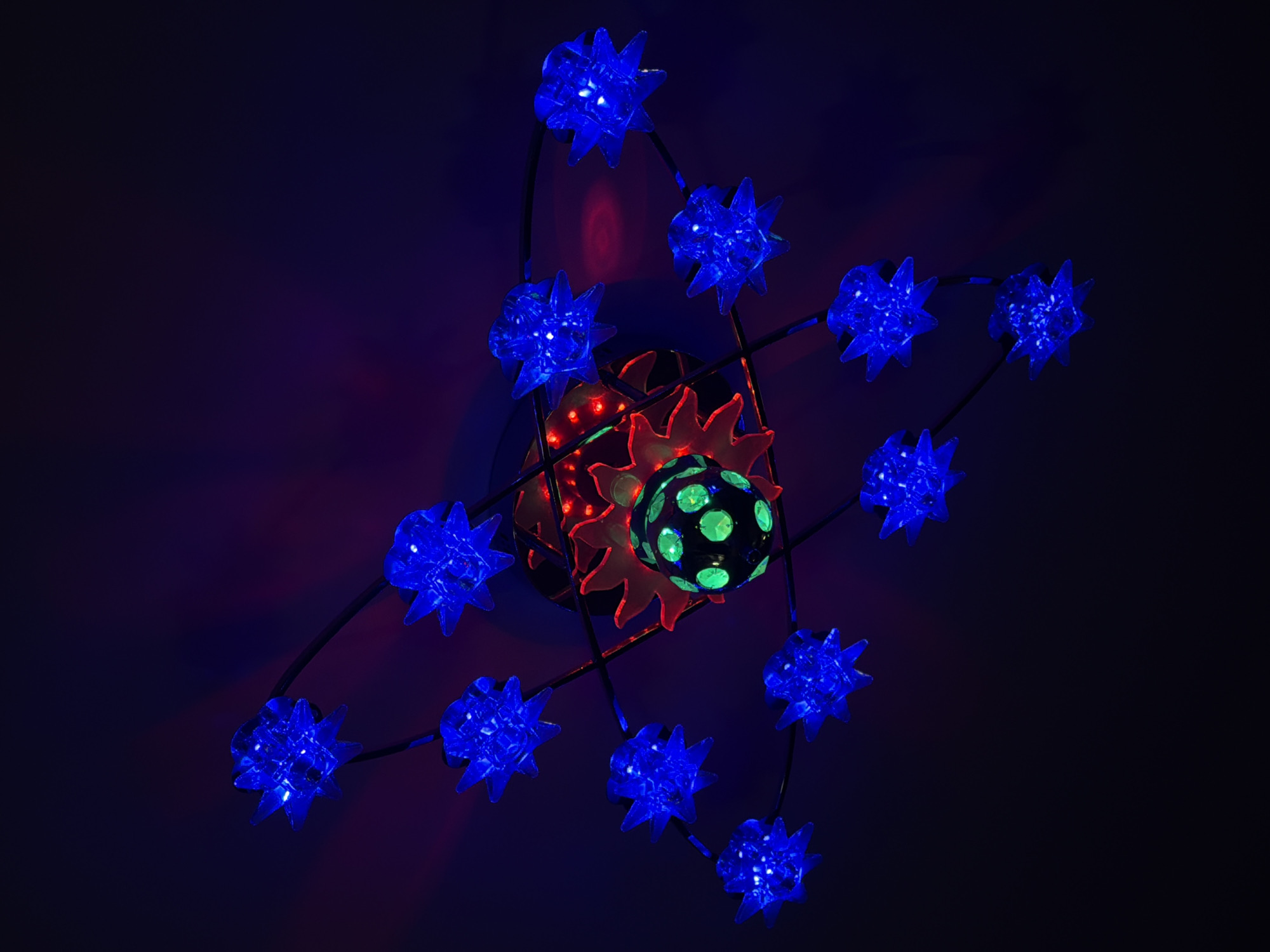 A photograph from an unknown location depicts a lighting fixture of unknown origin. The fixture consists of a central arrangement of red, orange, and green-speckled spheres surrounded by a series of twelve blue star-shaped lights connected by dark rods. The background is a gradient of dark blue and purple.