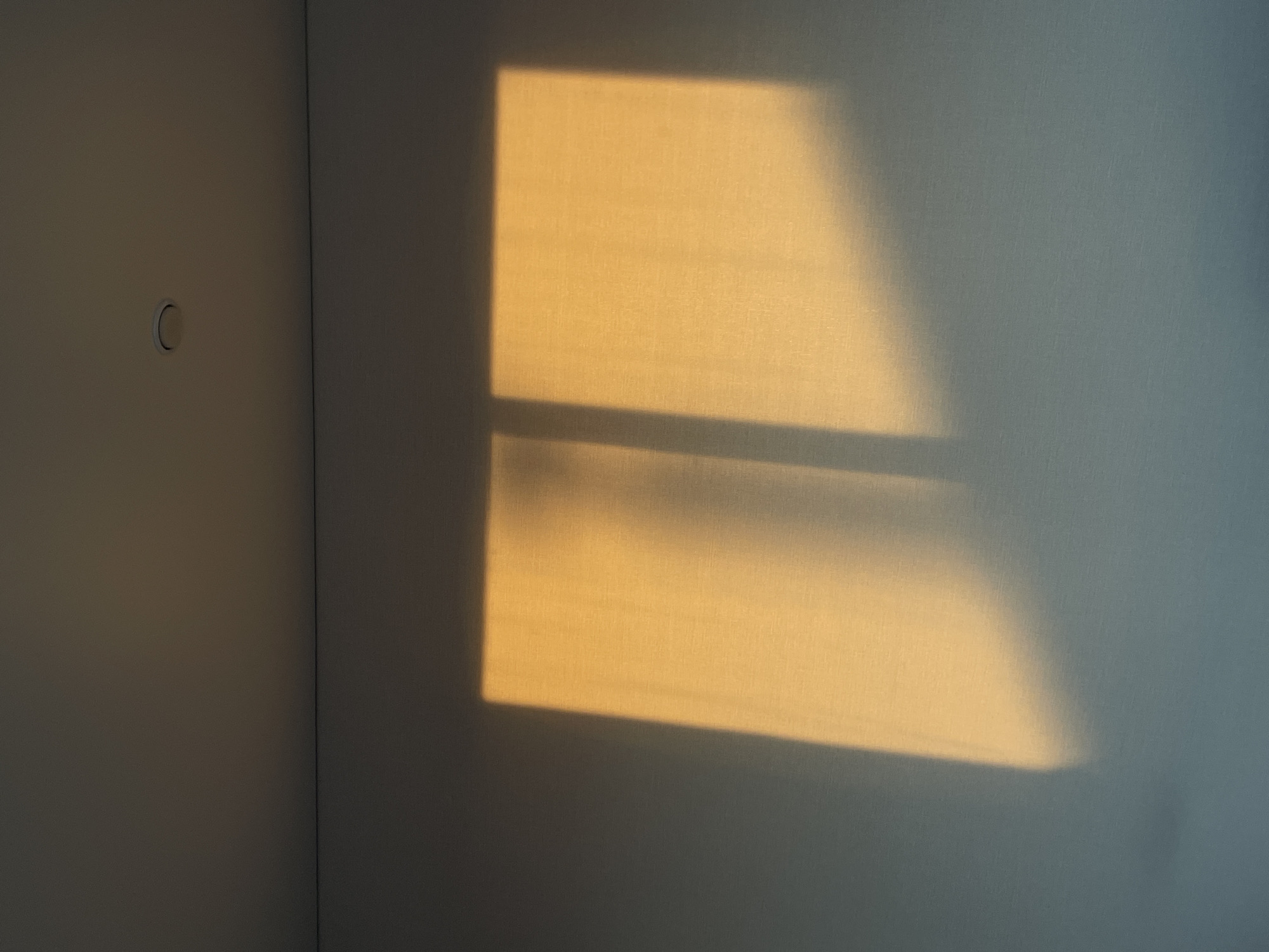 The image shows an interior corner of a room. The walls appear to be painted a light grey or off-white. A rectangular patch of golden sunlight is projected onto one of the walls, casting a shadow that suggests a window with a horizontal bar. On the adjacent wall, a circular object, possibly a light switch or cover plate, is visible. There is no indication of the location being in Подшивалово, Russia, or any other city, nor is there evidence of people, subjects, objects, actions, or interactions beyond the light and architectural details.