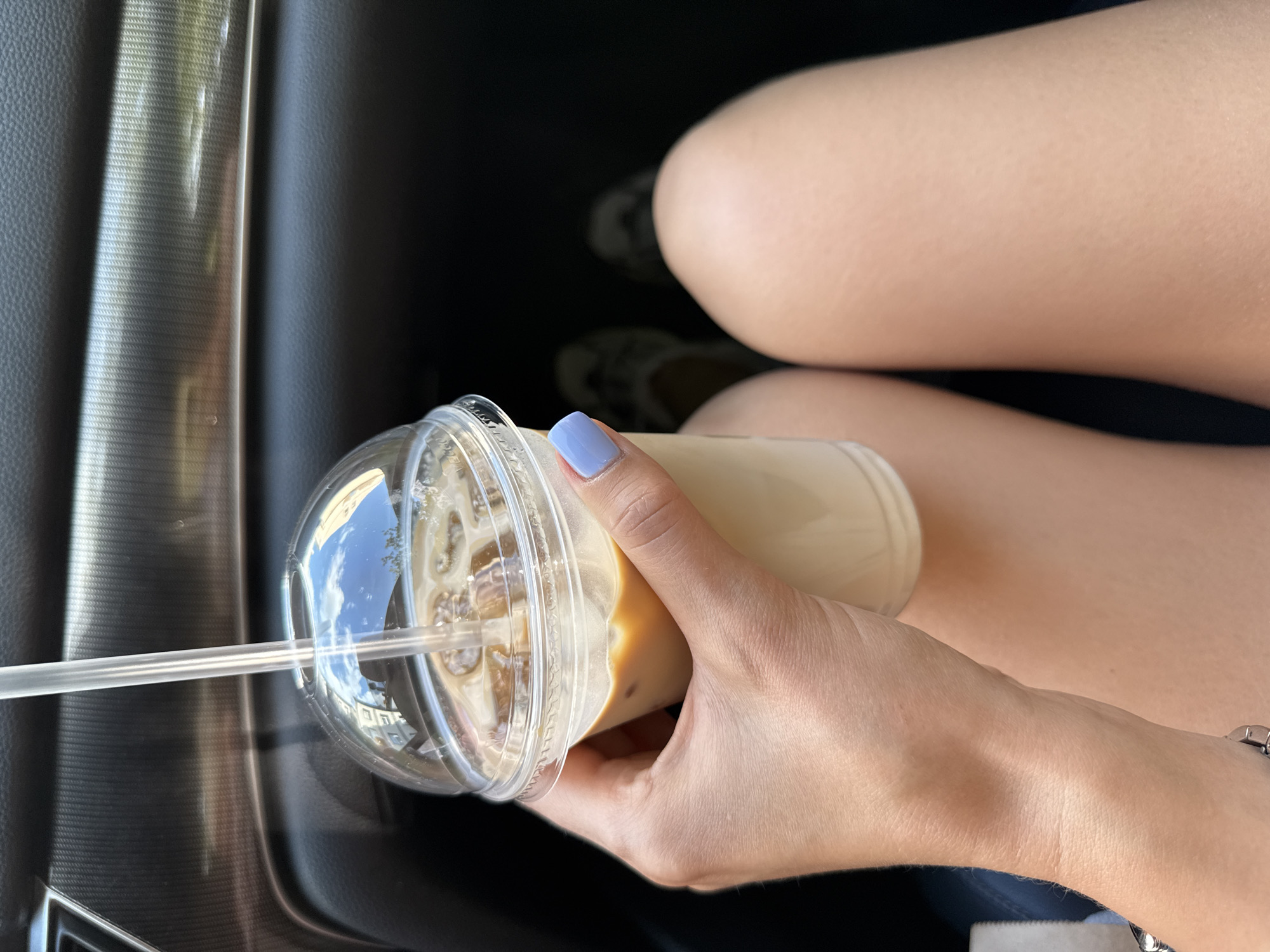 A person's hand, with light-colored skin and pale blue nail polish, holds a clear plastic cup filled with an iced coffee beverage. The cup has a domed lid with a clear plastic straw inserted. The beverage contains ice cubes and a light brown liquid. The background appears to be the interior of a car, with dark upholstery and a textured trim piece. In the lower portion of the image, a person's bare legs are visible, suggesting the presence of a female. The scene does not provide any identifiable information about the location within Mahilyow, Belarus.