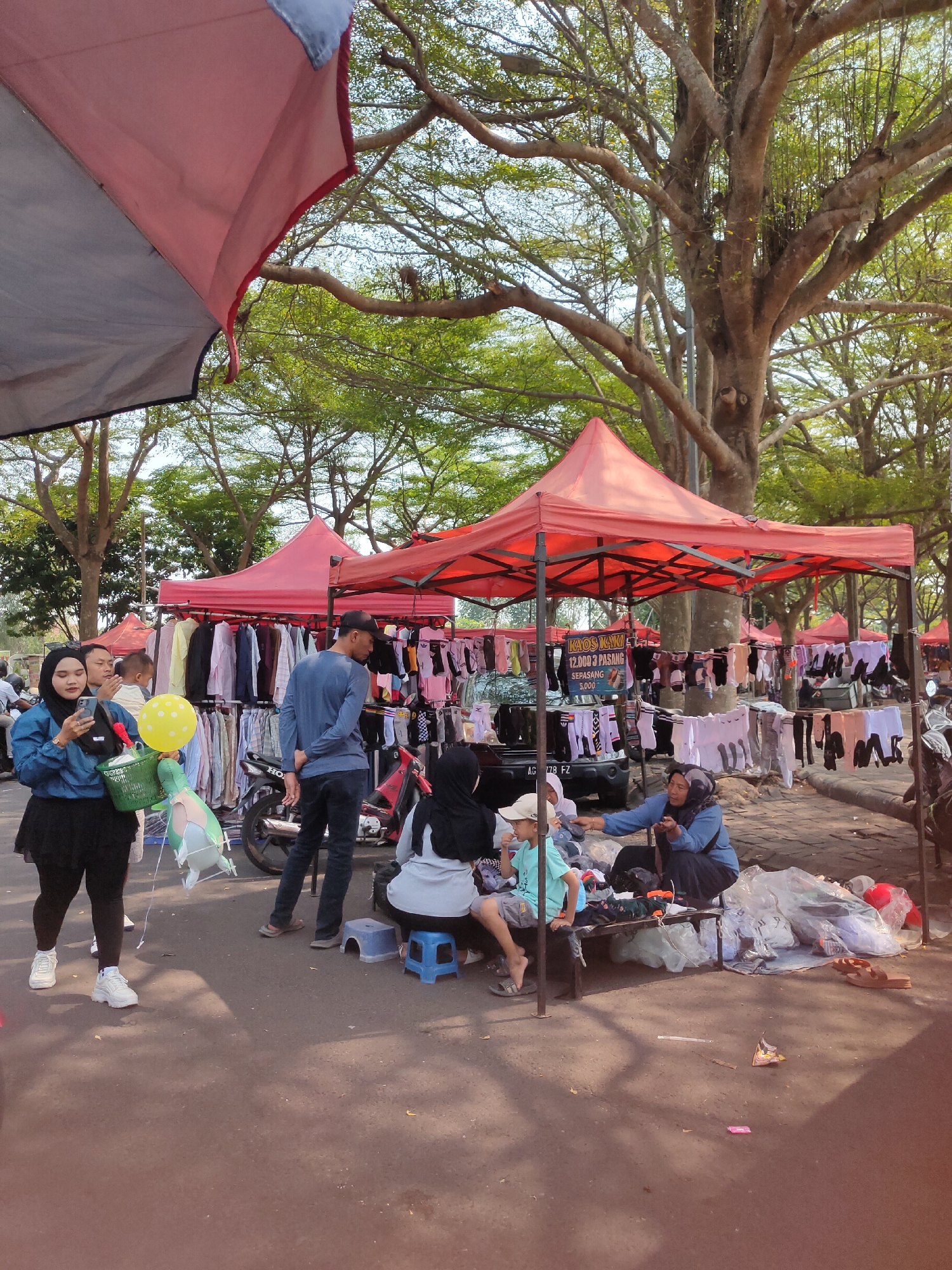 A busy outdoor market scene is depicted, likely in Ngasem, Indonesia, with several red canopy tents set up to display goods. In the foreground, on the left, a woman wearing a blue hijab and a blue jacket is holding a mobile phone and a green basket. Next to her, a child is holding a yellow balloon. Several people are browsing at the stalls. One stall prominently displays shirts on hangers, while another displays socks. A motorcycle is parked near one of the stalls. In the background, large trees with lush green leaves provide shade. The ground is a mix of asphalt and dirt. Some people are sitting on low stools or benches, engaged in conversation or shopping. Plastic bags and folded clothing are visible on the ground and on the benches, indicating the nature of the merchandise. The overall atmosphere suggests a vibrant local market day.