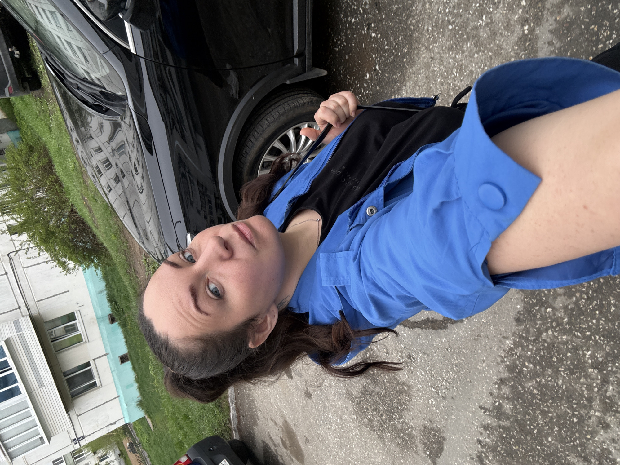 A woman is seen in a medium, slightly angled outdoor shot. She has fair skin, dark brown hair pulled back in a low ponytail, and blue eyes. She wears a black shirt under a blue button-down jacket.
She is positioned next to a black car, and behind her is an apartment building with windows. Green trees and grass are visible near the building. The ground is paved with asphalt, appearing slightly wet. The photo was taken in Chaikovsky, Russia.