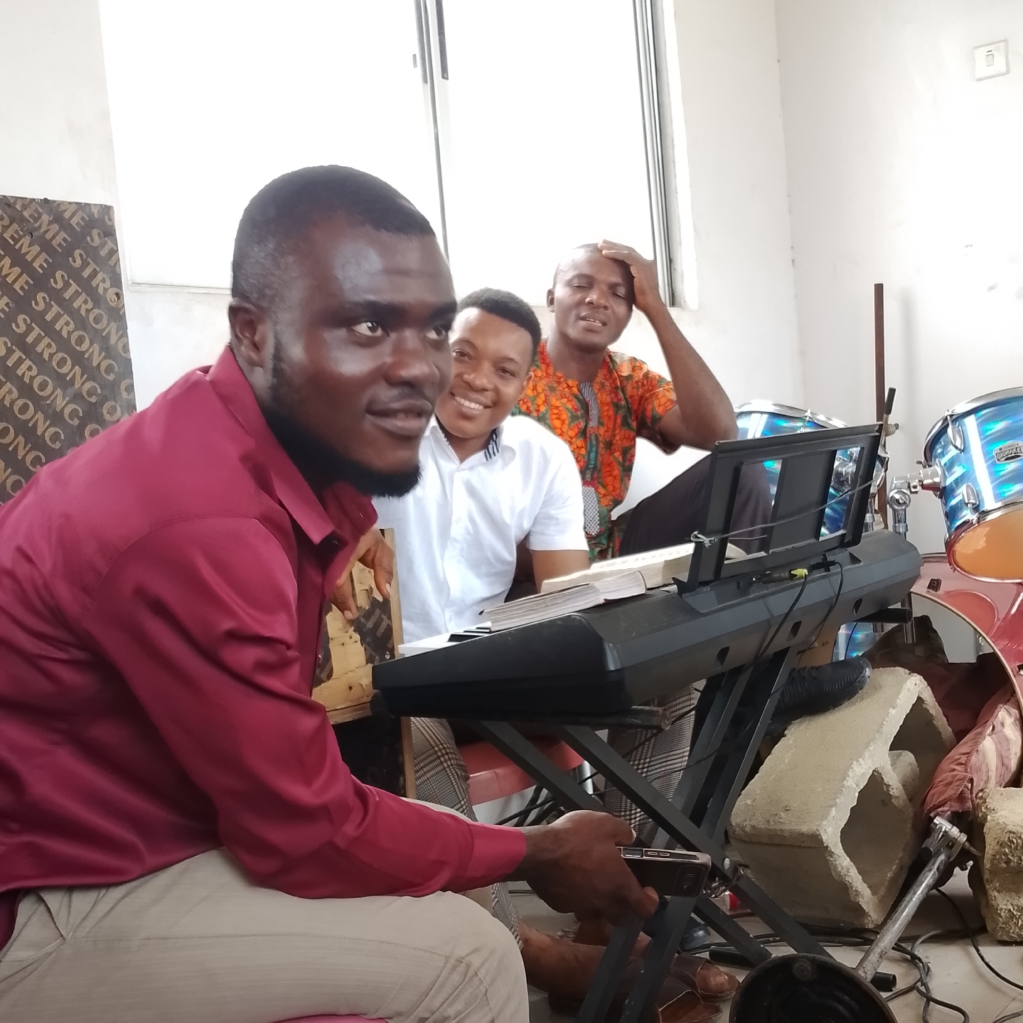 In a room in Iranla, Nigeria, three men are gathered around a keyboard and drums. The man in the foreground, wearing a burgundy shirt and beige pants, is looking towards the camera. To his right, a man in a white shirt smiles at the camera. Behind them, a third man in an orange and black patterned shirt rests his hand on his head. A keyboard with a music stand and sheet music is positioned in front of the men. Drums, one bright blue with white accents and another red, are visible in the background, suggesting a musical setting. The room appears to be a simple space with plain white walls and a window.