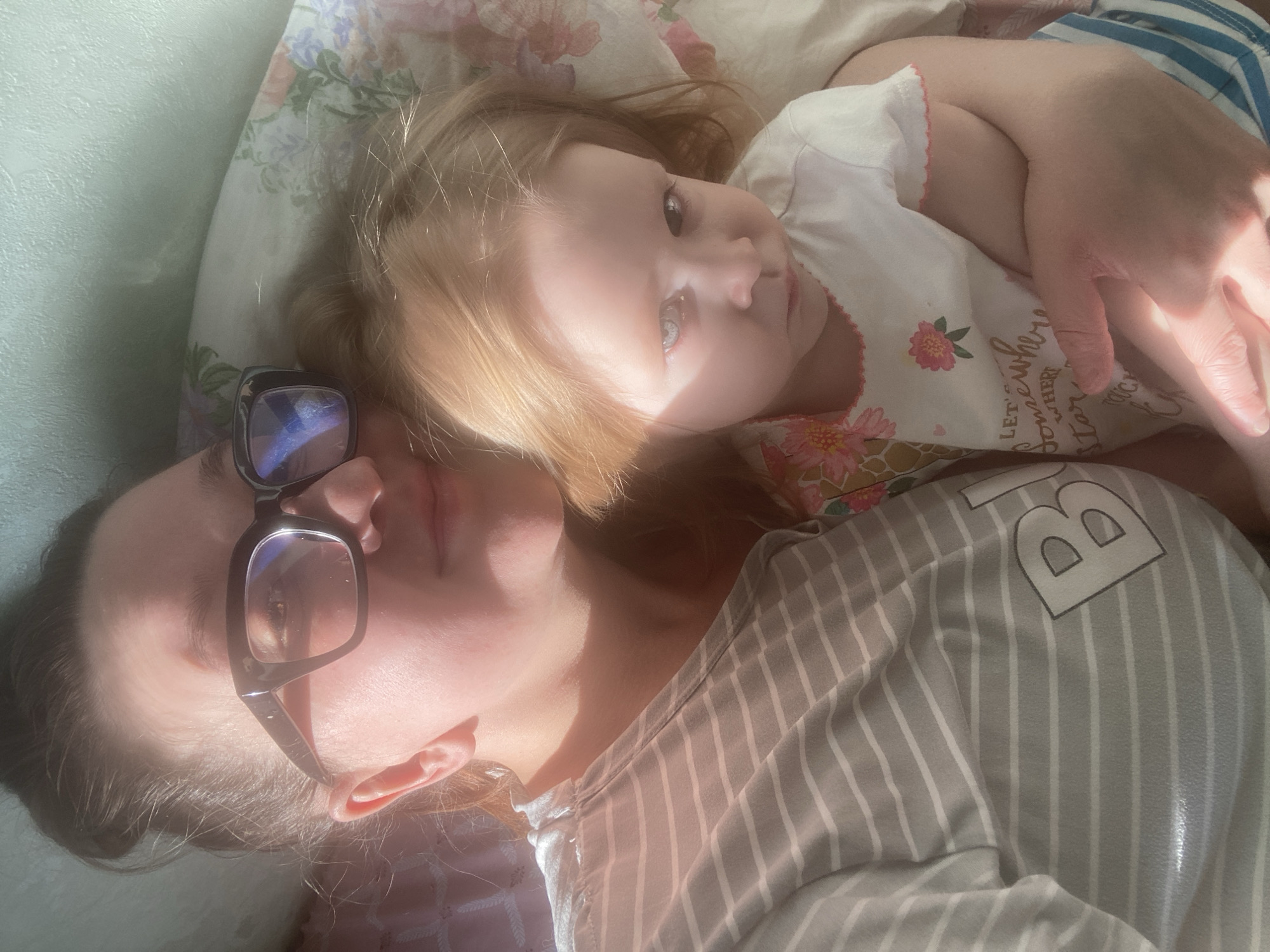 An adult female and a young child are depicted lying together indoors, presumably in a bed, under strong natural lighting. The adult female is positioned on the left, wearing black-framed glasses and a grey and white horizontally striped top. Her dark hair is pulled back, and her left arm embraces the child. The young child, with light reddish-blonde hair and light-colored eyes, is on the right, wearing a white t-shirt with a pink floral pattern and blue and white striped lower garments. The child's gaze is directed toward the viewer. Both subjects are resting on a pillow with a subtle floral pattern. A light-colored wall is visible in the upper background. No specific location within the city of Chaikovsky, Russia, is discernible from the visual evidence.