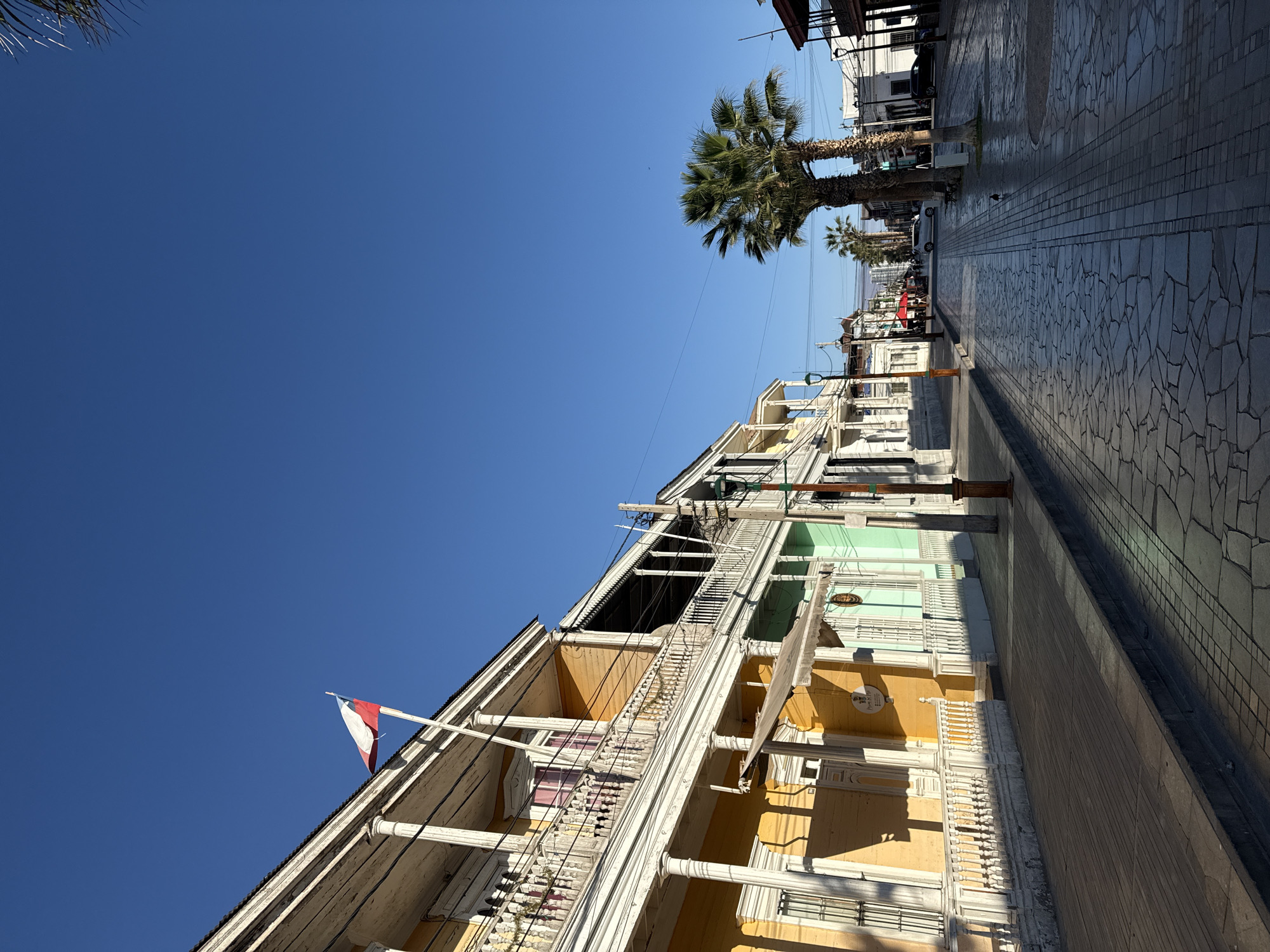 The image depicts a street scene in Iquique, Chile, characterized by colorful colonial-style buildings and a clear blue sky. The buildings, predominantly painted in yellow and green, feature ornate balconies with white railings and columns, typical of the region's architectural heritage. A Chilean flag flies from a pole on the balcony of a yellow building on the left. The street is paved with cobblestones and flagstones, reflecting the bright sunlight. Palm trees line the sidewalk, adding to the tropical ambiance. In the distance, there are more buildings, some with dark awnings, and a few vehicles are visible on the street. The scene suggests a quiet, sunny day in a historic part of the city.