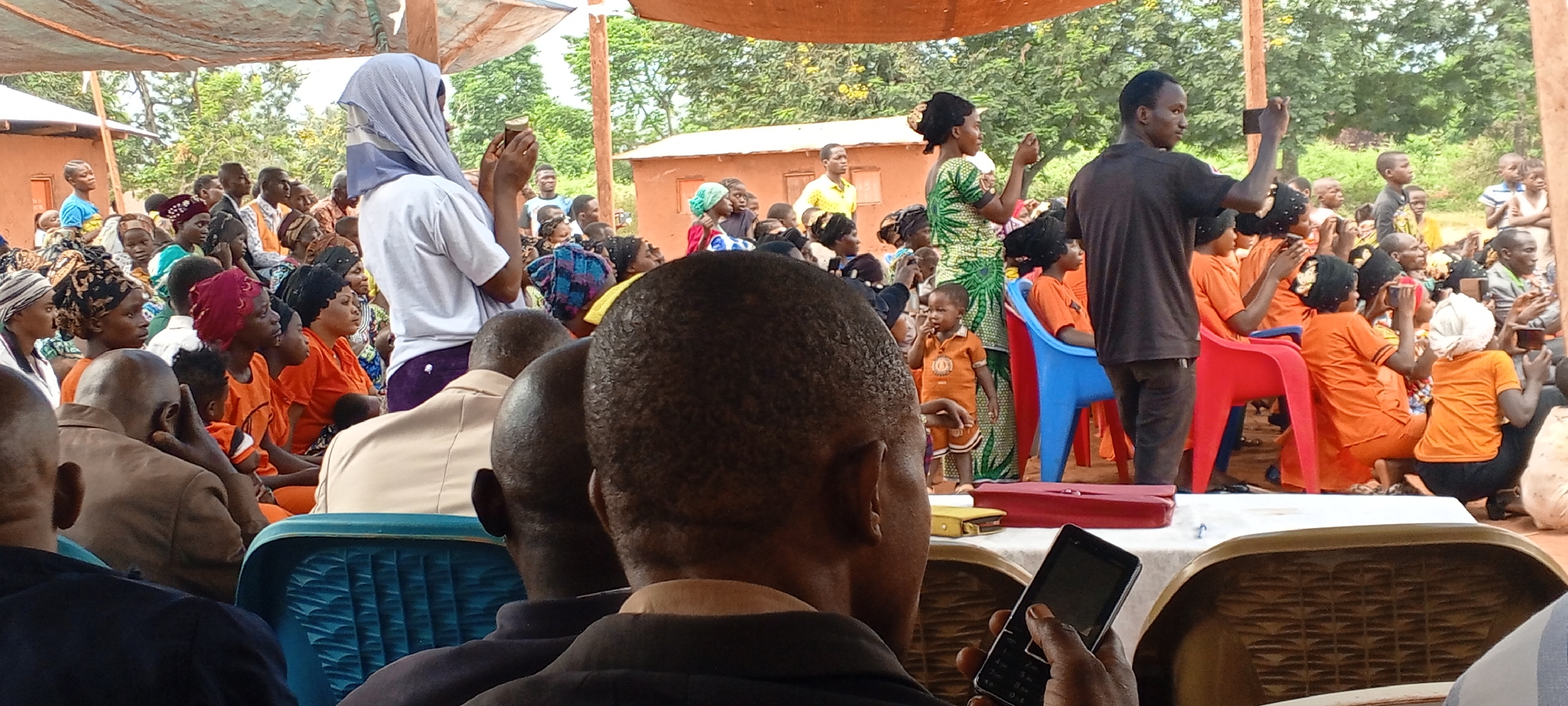 Captured in Ngalula, Democratic Republic of the Congo, a gathering unfolds outdoors under a tented structure. A seated crowd fills the foreground, observing individuals who are recording events on cell phones. Most of the crowd are dressed in orange, and there are several children among them. Other individuals at the forefront stand, and some are actively recording. A woman is wearing a green dress. In the midground, simple structures dot the landscape, with trees visible in the distance.