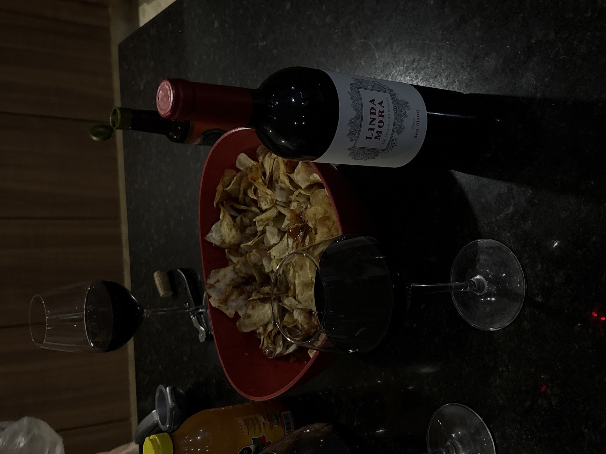 The scene is a dark countertop setting with a wine bottle labeled "Linda Mora De Finca Las Moras 2024 Red Blend". Next to it, there is an open bottle of wine and a red bowl filled with potato chips. Two wine glasses are filled with red wine. A bottle of juice and a wine opener are also present. The countertop is dark and speckled. The location in Santa Clara Ocoyucan, Mexico is not evident from the scene.