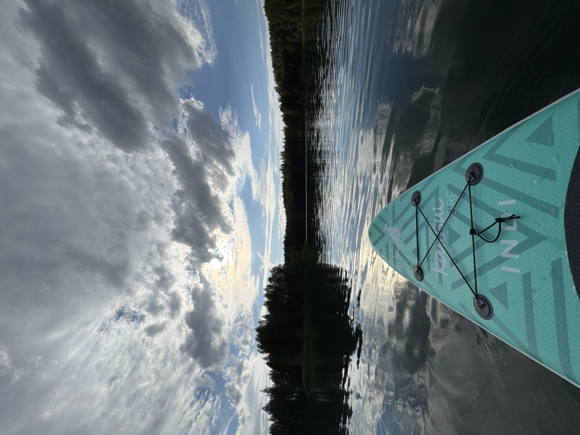 The image depicts a point-of-view shot from a paddleboard on a calm lake. The foreground features the front portion of a teal-colored paddleboard with a geometric pattern and black bungee cords. The water is dark and reflects the sky and the treeline on the distant shore. The treeline consists of dense, dark green trees, suggesting a forest. The sky is dramatic, filled with a mix of bright white and dark grey clouds, with patches of blue sky visible. A bright, hazy sun is visible through a break in the clouds towards the center-left of the frame. The overall impression is that of a serene, natural landscape, possibly in the area of Novyi, Russia, given its extensive natural areas and lakes. There are no people visible in the frame.