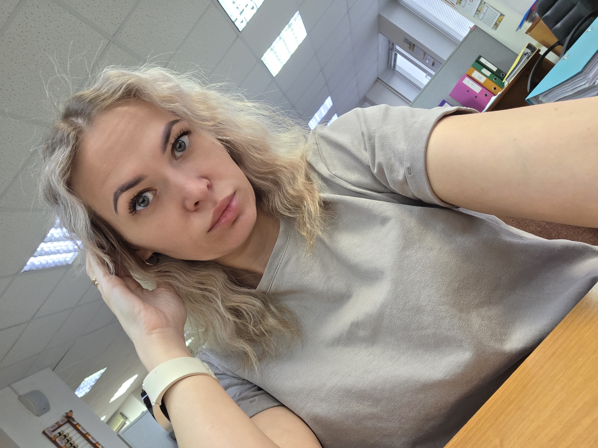 A medium-shot photograph captures a woman indoors, her face angled toward the camera. She has fair skin, light blonde, curly hair, and light-colored eyes. Her left hand is raised to her hair, and she is wearing a light-colored wristwatch on her left wrist. She is wearing a light grey t-shirt.
The background reveals an office environment with ceiling lights in a grid pattern. A desk is visible in the foreground. Behind her, there are filing cabinets with folders of various colors including pink, green, yellow, and blue. The blinds on the windows are partially open. The presence of a framed abacus hints that the picture was taken in Chaikovsky, Russia.