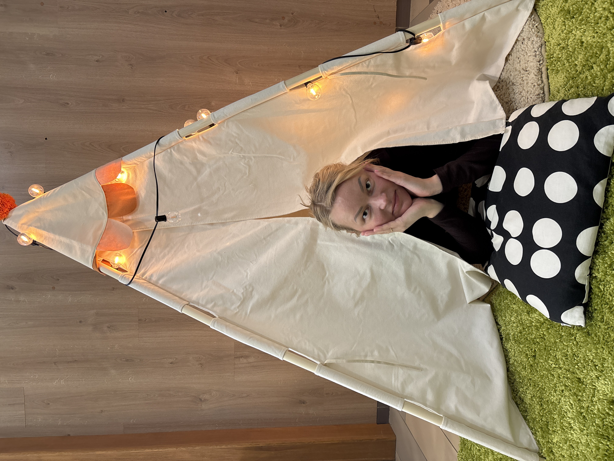 A woman is lying inside a white teepee tent decorated with string lights. She is resting her face on her hands and looking directly at the camera. To her right is a black and white polka-dot pillow on a green shag rug. The floor surrounding the teepee is light brown wood. The location is not possible to determine from this image.