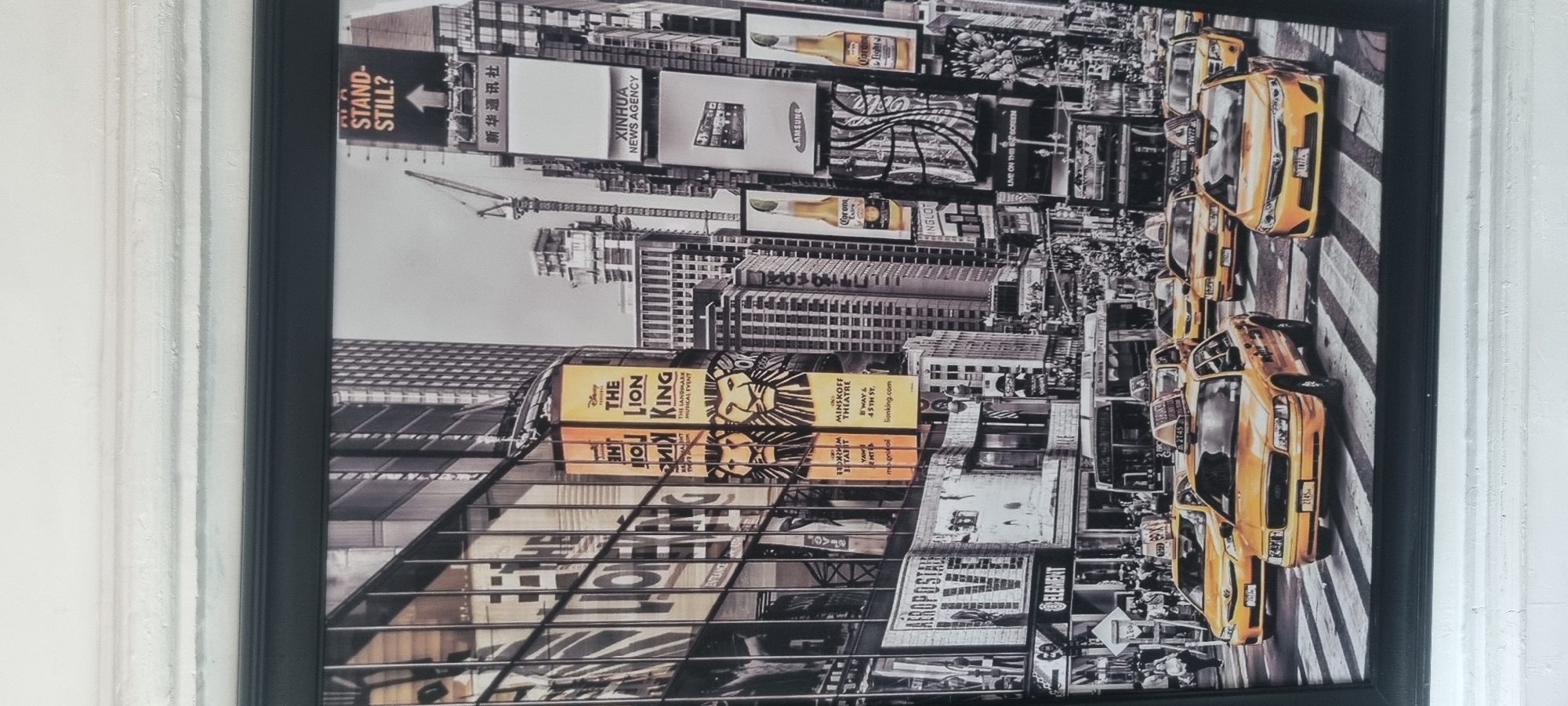 The image displays a bustling urban street scene, rendered primarily in black and white with selective colorization. The scene is dominated by towering buildings adorned with numerous large digital billboards and advertisements, characteristic of a major commercial hub. A prominent line of yellow taxis occupies the street in the foreground and midground, their iconic color standing out vividly against the monochromatic backdrop of the surrounding city. These taxis appear to be in traffic or waiting at an intersection marked with a crosswalk.
Visible text on the billboards includes "THE LION KING" musical, explicitly stating "MINSKOFF THEATRE" and the address "B'WAY & 45TH ST.", which clearly indicates Times Square, New York City. Other advertisements feature brands such as "XINHUA NEWS AGENCY", "SAMSUNG", "Corona", "INGLOT", "AEROPOSTALE", and "ELEMENT". A construction crane is visible among the skyscrapers in the background. The time of day appears to be daytime, suggested by the brightness and lack of heavy artificial illumination dominating the scene. The image conveys the energetic and vibrant atmosphere of a busy metropolitan area.