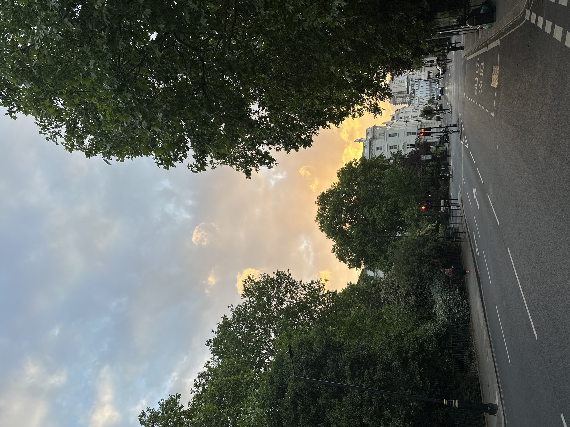The image depicts a street scene in London, United Kingdom, likely taken during the late afternoon or early evening as suggested by the golden hue of the sky. The view is oriented down a wide street lined with trees, which partially obscure the buildings along the road. The architecture consists of white buildings, some of which appear to be apartment complexes or hotels. The sky above is partially cloudy with patches of golden light indicating the setting sun. The street itself is asphalt and features a bus stop marked with white paint. A few pedestrians and vehicles can be seen in the distance, and the street is lined with trees.