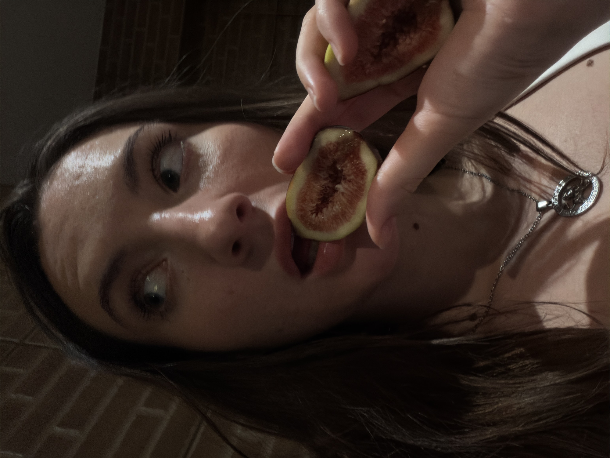 The image shows a close-up of a young woman holding a halved fig to her mouth, with the other half of the fig in her hand. She has dark brown hair and is looking to the side. Her lips are slightly parted as if about to take a bite. She is wearing a white top and a silver necklace. The background appears to be a brick wall, which is common in many urban settings. The lighting suggests an indoor environment, possibly at night, given the shadows. There is no visual information in the image that would allow for the identification of the location as Sharm El Sheikh, Egypt.