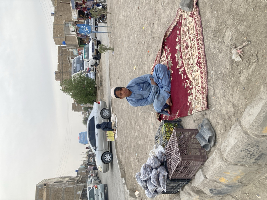 The image displays a street scene in Taftan, Pakistan, under an overcast sky. In the foreground, a male child, approximately 8-12 years old, with dark hair and a light complexion, is seated barefoot and cross-legged on a partially unfurled red patterned rug. He is wearing a light blue shalwar kameez and a white skullcap, looking directly forward. To the child's left, on a dirt patch bordered by a concrete curb with yellow markings, three plastic crates are positioned. Two dark brown crates are stacked, containing transparent plastic bags filled with a dark, possibly dried, food product. A green crate holds green produce. A pair of grey open-toed sandals rests on the dirt beside the crates. The ground surrounding the rug is dusty, with minor debris visible.
The midground reveals a paved street with vehicle and pedestrian activity. A silver sedan is parked or moving slowly. Behind it, another person, dressed in dark attire, is seated on the ground, attending to multiple yellow plastic containers placed on a white cloth. Further down the street, a large white passenger bus with "HASNAIN" visible on its front, along with a white pickup truck and other vehicles, are present. A blue street sign with white Arabic script stands on the right side of the road. Buildings with flat roofs and storefronts line both sides of the street. Overhead power lines are visible. Small trees and bushes are scattered within the dusty environment. The background shows additional urban structures extending towards the horizon under the muted sky.